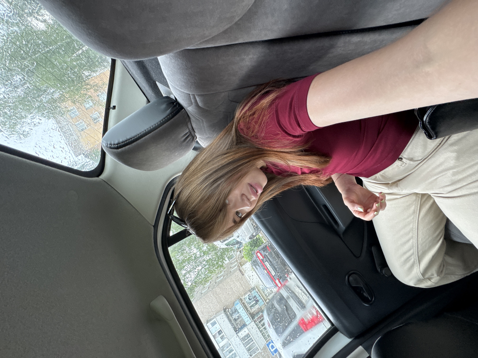 A young woman with long, light brown hair and bangs is sitting in the back seat of a car, looking towards the viewer. She is wearing a dark red t-shirt and light beige pants. Her right arm is extended towards the camera, and her left hand is resting on her lap. The car appears to be parked on a street in Chaikovsky, Russia, as indicated by the license plate on a car visible through the window. It is raining, with raindrops visible on the car windows and a building and trees in the background. The interior of the car is visible, including the grey fabric seats and the car door.