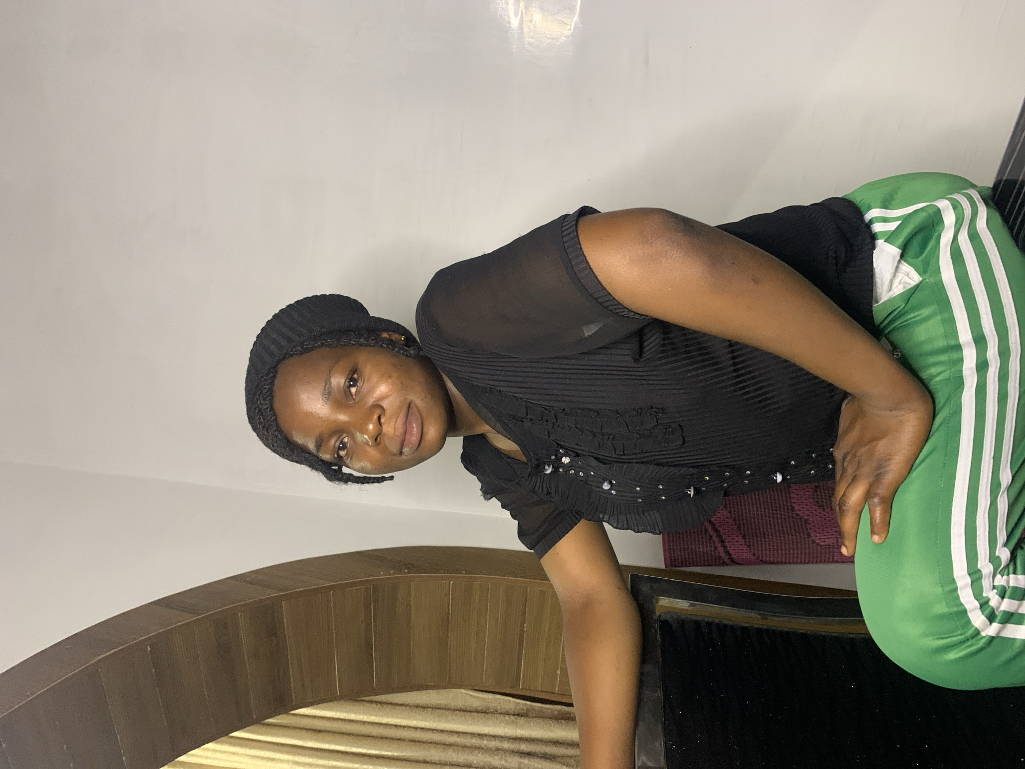 A young Black woman wearing a black beanie, a black sheer top with ruffles and buttons, and green track pants with white stripes, sits facing forward and slightly to the right. She is leaning on a dark chair or table. Behind her, to the left, is a light-colored curtain partially visible through a wooden archway. The background wall is white, with a faint reflection on the right side. The scene appears to be indoors. The location is not specified in the image, and therefore cannot be determined to be Jos, Nigeria.