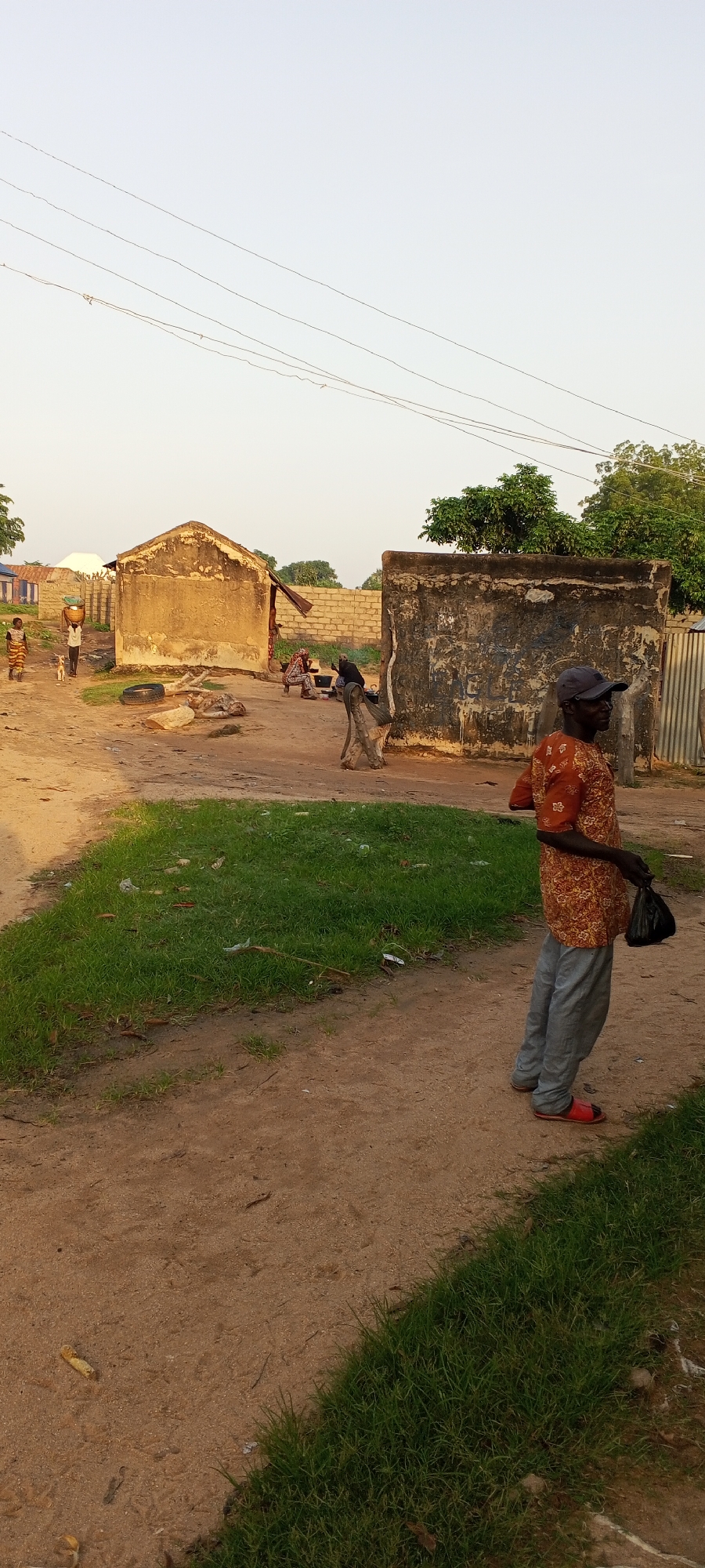 A man stands on a dirt path in Jalingo, Nigeria, holding a black plastic bag in his right hand. He is wearing a red and orange patterned shirt, grey pants, and red sandals. Behind him, a rustic, weathered concrete structure with peeling paint stands to the right, and a smaller, dilapidated mud-brick building with a corrugated metal roof is to the left. Several other people are visible in the background, including a person carrying a basket on their head. The environment is characterized by unpaved ground, sparse vegetation, and overhead power lines, suggesting a rural or semi-urban setting.