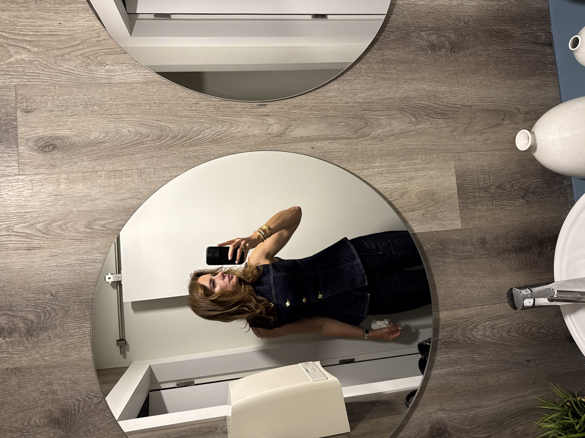 The image depicts a woman taking a selfie in a bathroom with two circular mirrors. The bathroom floor is covered in wood-grain planks. Reflected in the lower mirror is the woman holding a phone. She is wearing a dark denim vest with gold buttons, dark pants, and jewelry on her wrists. The reflection also reveals a white vanity and a white structure on top of it, possibly a toilet or a seat. To the right of the mirror's edge are a white sink, a silver faucet, and decorative white vases. The scene appears to be indoors. The location is Lisbon, Portugal.