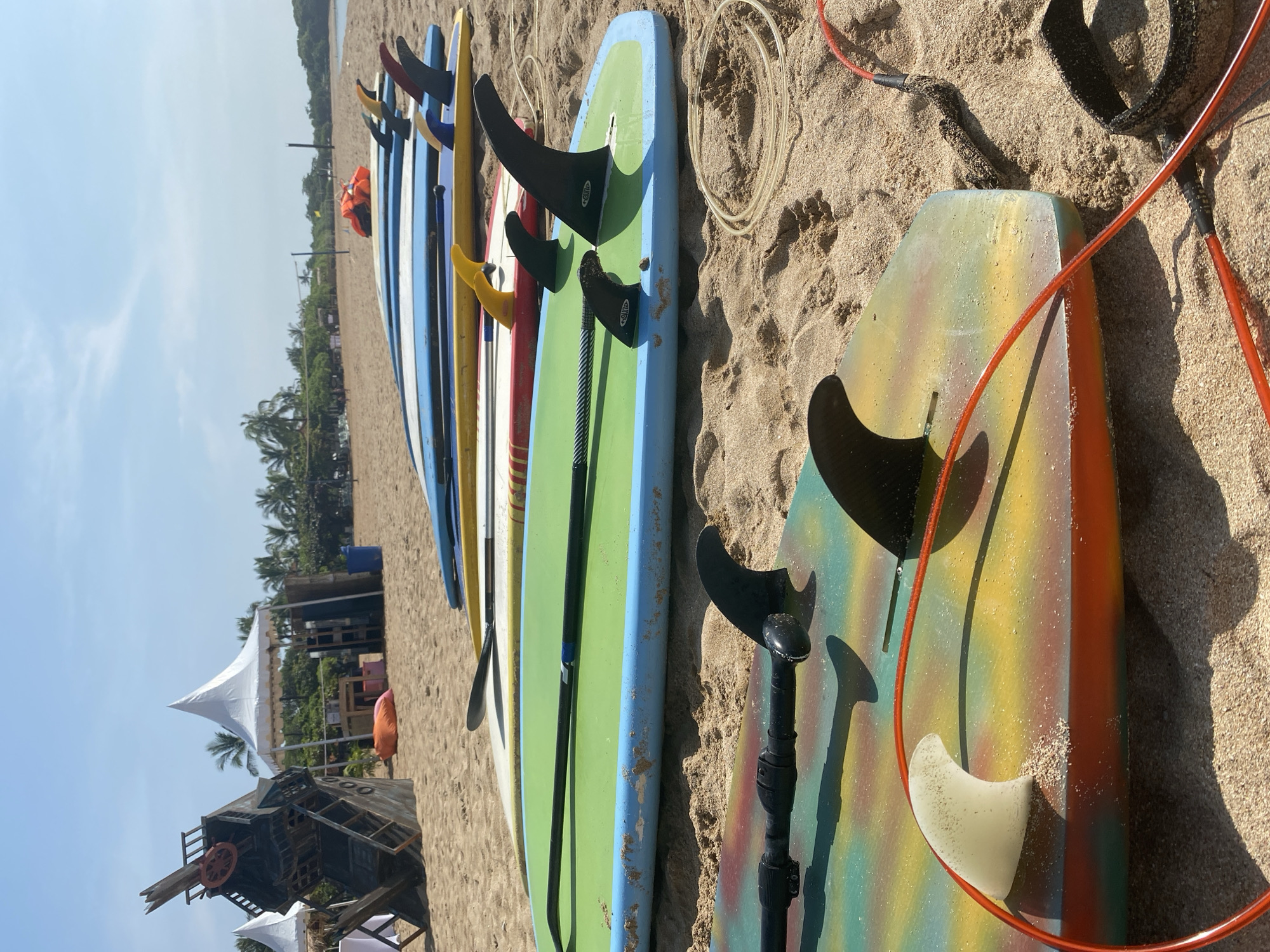 The image captures a row of surfboards and paddleboards lined up on a sandy beach in Nusa Dua, Indonesia. The boards are of varying colors and designs, with fins and paddles visible. In the background, there are trees, beach structures including a windmill decoration, and a tent structure. The sky is clear and blue. The boards rest directly on the sand, and the scene appears to be a rental or storage area for watersports equipment. The scene appears to be set during daytime with ample sunlight.