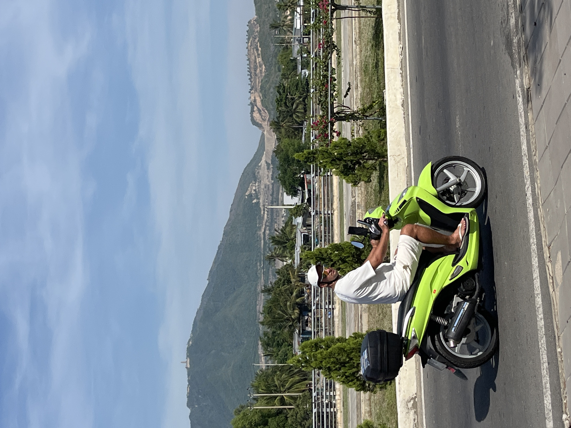A man wearing a white t-shirt, white shorts, a white baseball cap, and sunglasses rides a lime green scooter on a paved road. The scooter has a black storage box attached to the rear. In the background, there is a lush green mountain range with some visible construction or mining activity on one of the slopes. A tall monument or structure is visible at the summit of the highest peak. A line of trees and buildings are situated at the base of the mountains, with a metal fence and more trees along the roadside. The sky is blue with scattered white clouds. The scene appears to be in a coastal or hilly area of Nam Nha Trang Ward, Vietnam, given the distinctive mountainous landscape and tropical vegetation.