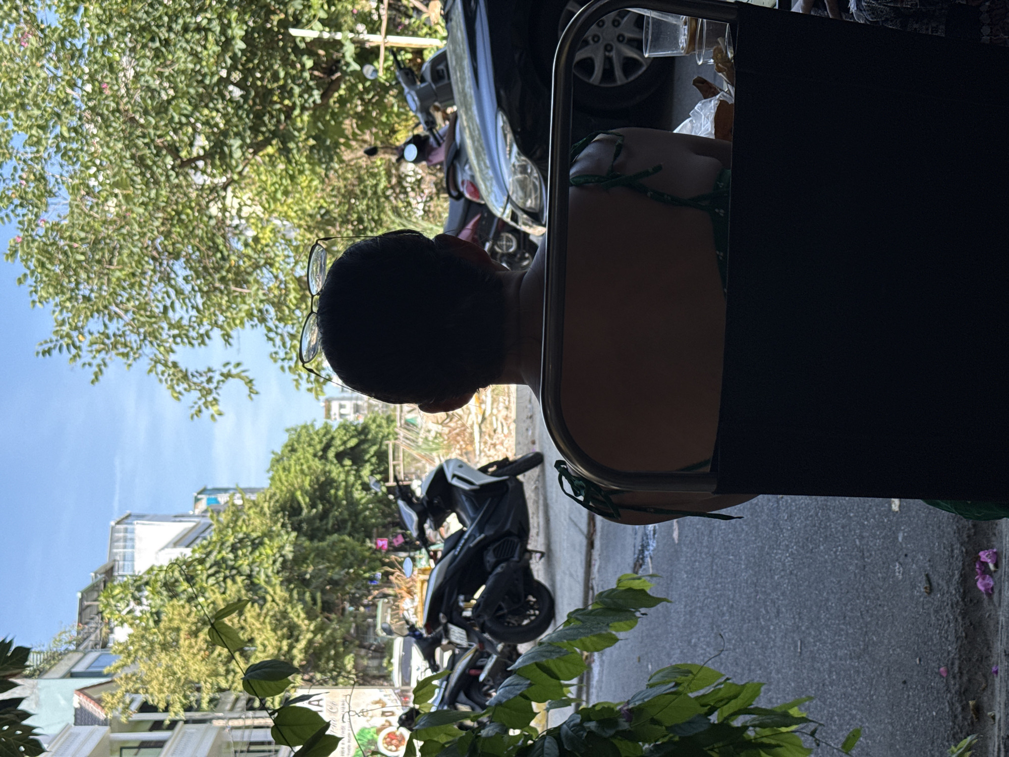The image shows a person sitting in a cafe or outdoor seating area, facing away from the camera. They are wearing a dark top with thin green straps. A pair of glasses rests on top of their head. The person is seated at a table, and part of a black chair is visible in the foreground.
In the background, a street scene unfolds. Several motorcycles are parked along the sidewalk, including a white and black scooter. A black car is also visible. Lush green trees and buildings line the street, suggesting an urban environment. The sky is clear and blue. Fallen pink petals are scattered on the pavement near the seating area.
This scene is consistent with a street-side cafe in Đà Nẵng, Vietnam, a city known for its vibrant street life, cafes, and abundance of motorcycles. The architecture and vegetation are also characteristic of Southeast Asian cities.