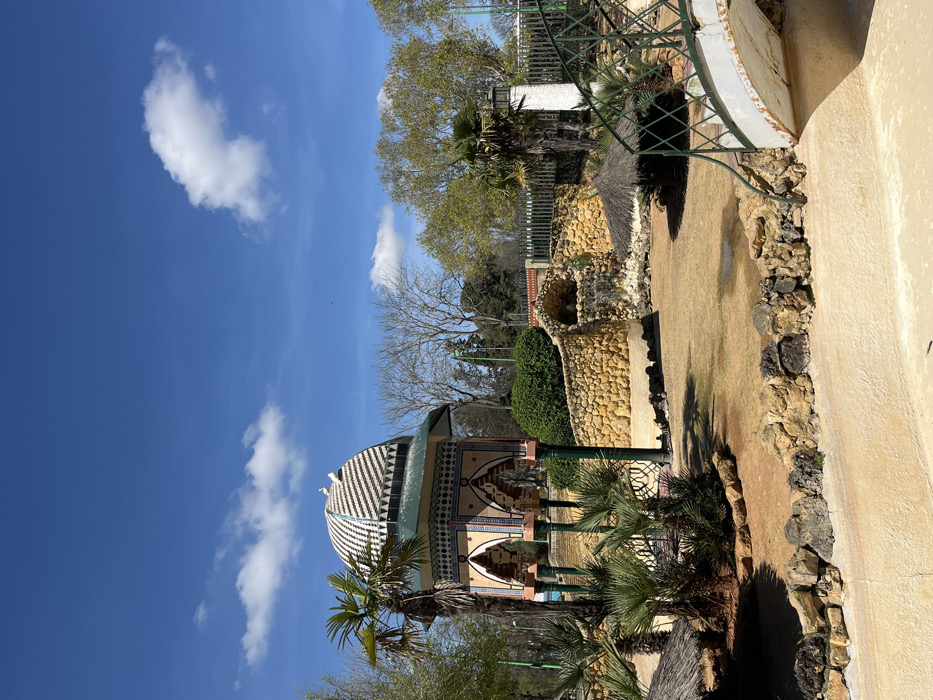 The image depicts a section of Parque de La Alquería in Dos Hermanas, Spain, under a clear blue sky with scattered white cumulus clouds. The scene is illuminated by strong daylight, casting distinct shadows.
The central subject is a multi-sided pavilion or gazebo of Moorish architectural design, situated slightly left of center. It features a domed roof with a black and white geometric checkered pattern, upon which two white birds are perched. Below the dome, the structure has a teal-green base and arched openings supported by slender columns, all adorned with intricate mosaic-like patterns in blue, orange, and light brown hues.
In the mid-ground, the terrain is primarily dry, light brown earth or sparse, dry grass, with several small pools of standing water. Various palm trees are distributed throughout, along with patches of dense green shrubs and hedges. To the right of the pavilion, a man-made rock formation includes a small waterfall cascading into a lower basin. Behind these features, deciduous trees with bare branches are visible, alongside trees with full green foliage.
The immediate foreground consists of a curved, light-colored concrete or stucco barrier. On the right side of the foreground, a decorative green metal bridge with white, arching supports is partially visible, extending from the lower right corner towards the center. In the background, a green metal fence can be seen beyond the trees. No human figures are present in the frame.