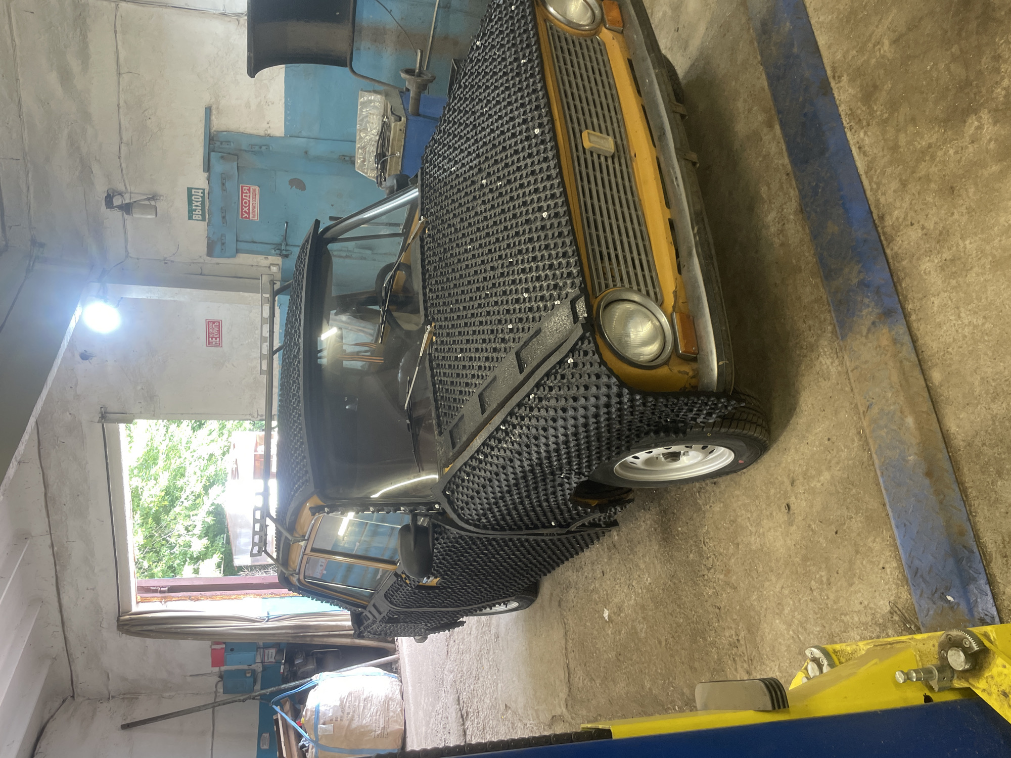 A modified yellow sedan, possibly a Lada, is parked inside a garage in Laishevo, Russia. The car's exterior, including the hood and sides, is covered in a black textured material with numerous small protrusions, resembling a form of automotive armor or protection. The car also has a roof rack. The garage appears to be a workshop, with tools, machinery, and a large bag of material visible in the background. A blue door with Russian signage indicating "EXIT" and another sign about "CARE" is present on the back wall. The floor is concrete, and a yellow car lift is positioned in the foreground. The scene is lit by overhead lighting and daylight streaming through an open doorway.