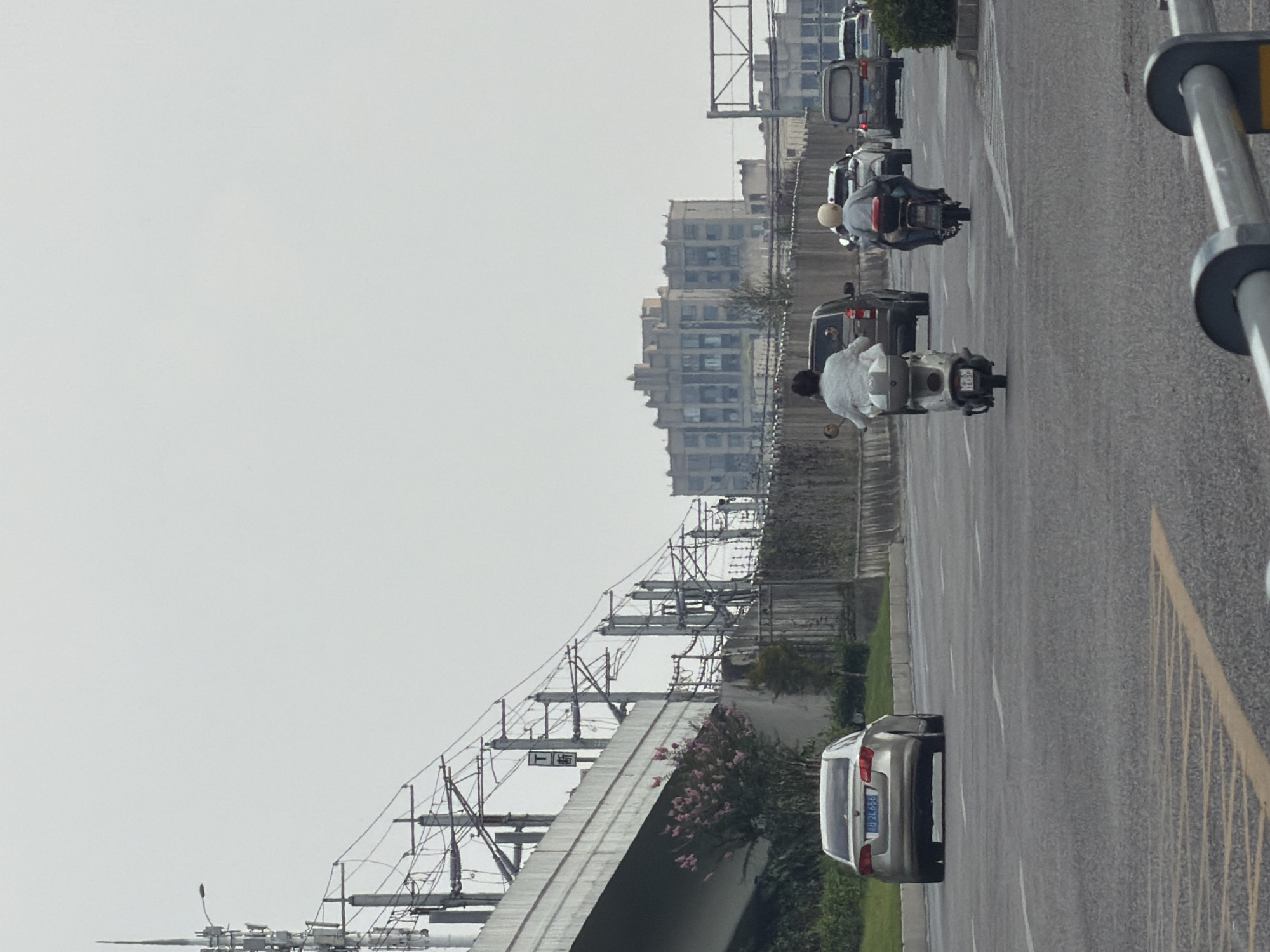 The image captures a view of a road with several vehicles, including cars and motorcycles, in Jiangjin, China. In the foreground, a silver sedan is visible on the right side of the road. Further down the road, two motorcycles are traveling in the same direction. The first motorcycle, on the left, has a person wearing a white jacket. The second motorcycle, to the right, has a person wearing a blue shirt. Behind them, several cars and a van are also present on the road. Overhead, a complex network of power lines and a highway overpass suggest an urban or suburban environment with significant infrastructure. In the background, a large, multi-story building with a distinctive architectural style stands against a pale sky. The overall scene depicts a traffic flow on a relatively overcast day.