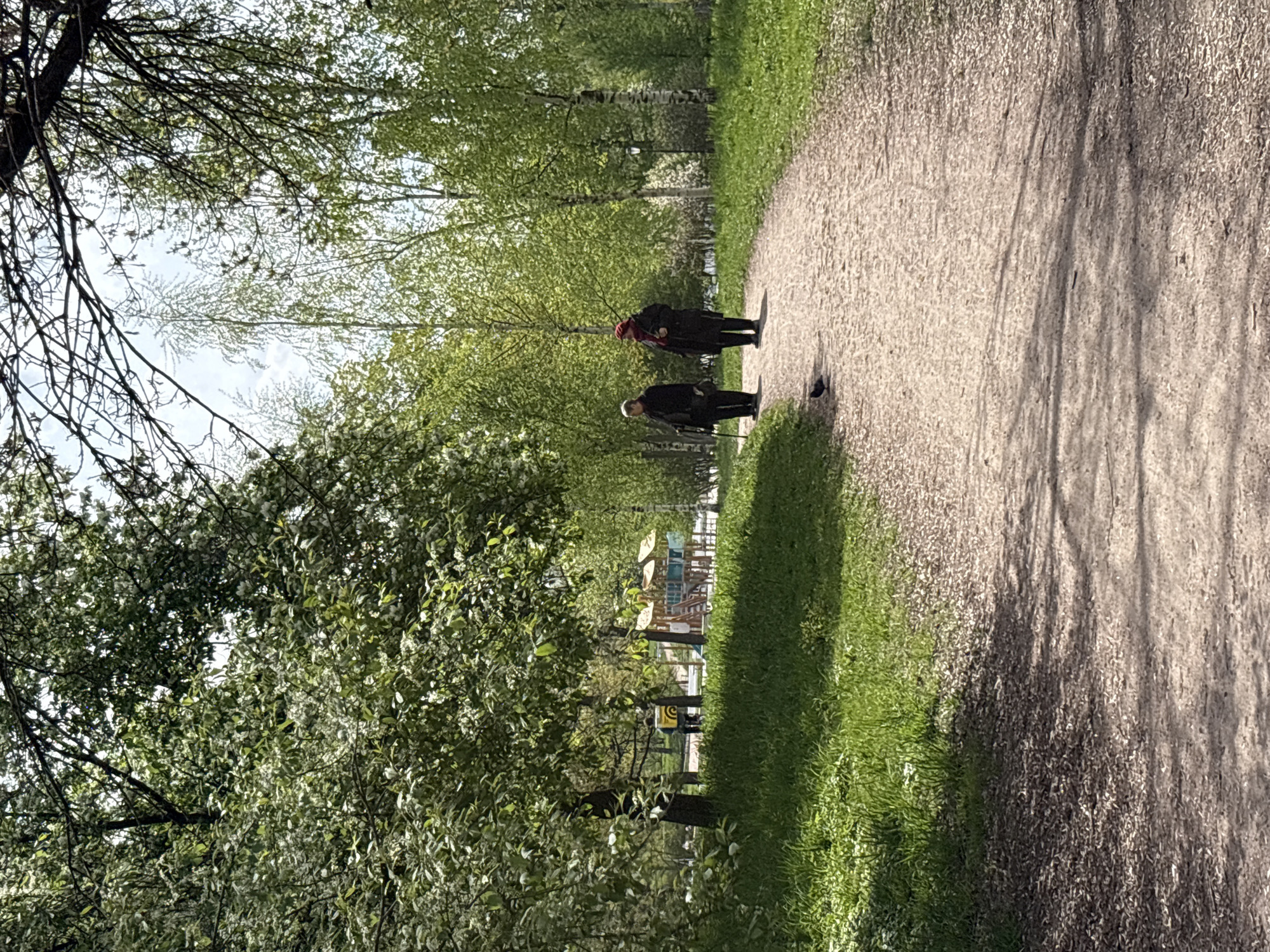 The image depicts a gravel pathway in a park-like setting. Two individuals are visible on the pathway. One person is wearing dark clothing and a red head covering; the other is wearing dark clothing. Trees and green foliage line the pathway. In the background, playground equipment is discernible. The location is potentially in Saint Petersburg, Russia.