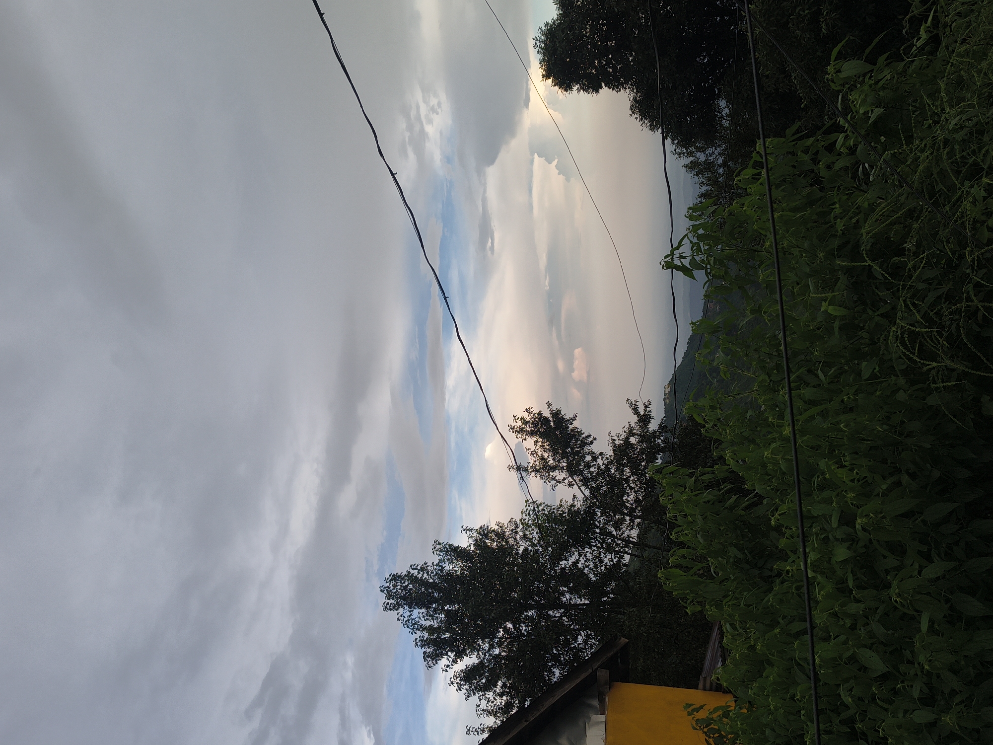 The image depicts a cloudy sky above a hilly landscape, possibly Haini, India. A bright yellow structure, likely a building, is visible on the left, partially obscured by dark green foliage. A large, dark tree with dense leaves stands to the right of the building. The sky is filled with a mix of gray and lighter, patchy clouds, with hints of pastel colors suggesting either sunrise or sunset. Power lines stretch horizontally across the upper portion of the frame. In the distance, the hazy outlines of hills and possibly some human settlements are discernible. The foreground is dominated by lush, green bushes and vegetation.