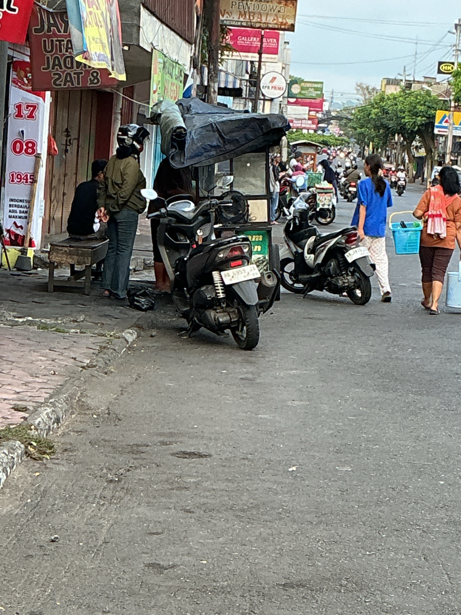 The image captures a street scene in Magelang, Indonesia, likely during the day, with a focus on the activity in front of a shop. The foreground is dominated by an asphalt road and a paved sidewalk. Several motorcycles are parked or in motion, indicating a busy urban environment.
On the left side of the image, a shop displays red and yellow banners with Indonesian text, one prominently showing "17 08 1945". Two individuals are present near the shop; one, wearing a dark shirt and seated on a wooden bench, and another, in a green jacket and helmet, standing. A dark tarp covers an object behind them, possibly a food stall or a cart.
Further down the street, the scene opens up to a bustling road filled with numerous motorcycles, suggesting traffic. Shops and signs are visible along the street, some displaying corporate logos and advertisements. Two women are walking away from the viewer towards the right side of the frame; one is wearing a blue shirt and white pants, carrying a blue basket, and the other is in an orange top and brown pants, also carrying a basket. The overall impression is of a lively, commercial area in an Indonesian city.