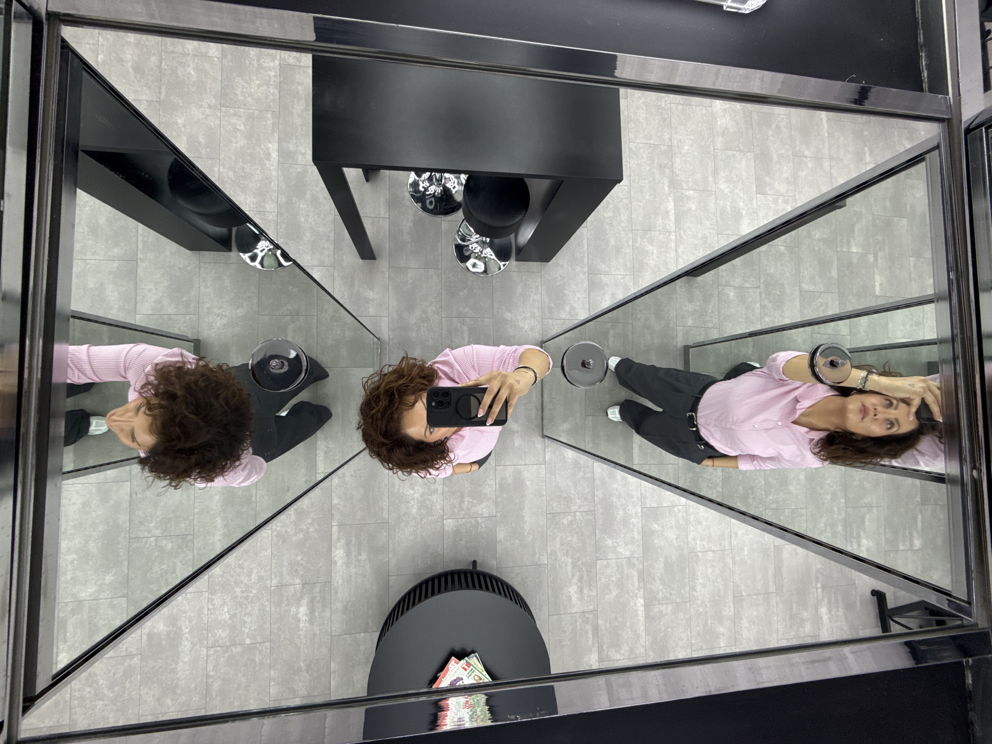 A high-angle shot depicts a woman standing within a mirrored enclosure. She holds a smartphone, presumably taking the photograph. The mirrors create a kaleidoscopic effect, reflecting the woman and the surrounding environment multiple times. The woman has curly brown hair and is wearing a pink striped shirt and dark pants. There is a black table with chairs and a round table with what appears to be money on it in the shot. The floor is tiled with grey stone and has a neutral aesthetic. The location appears to be a shop of some sort, but the specific location in the city of Unknown, Unknown is not discernible.