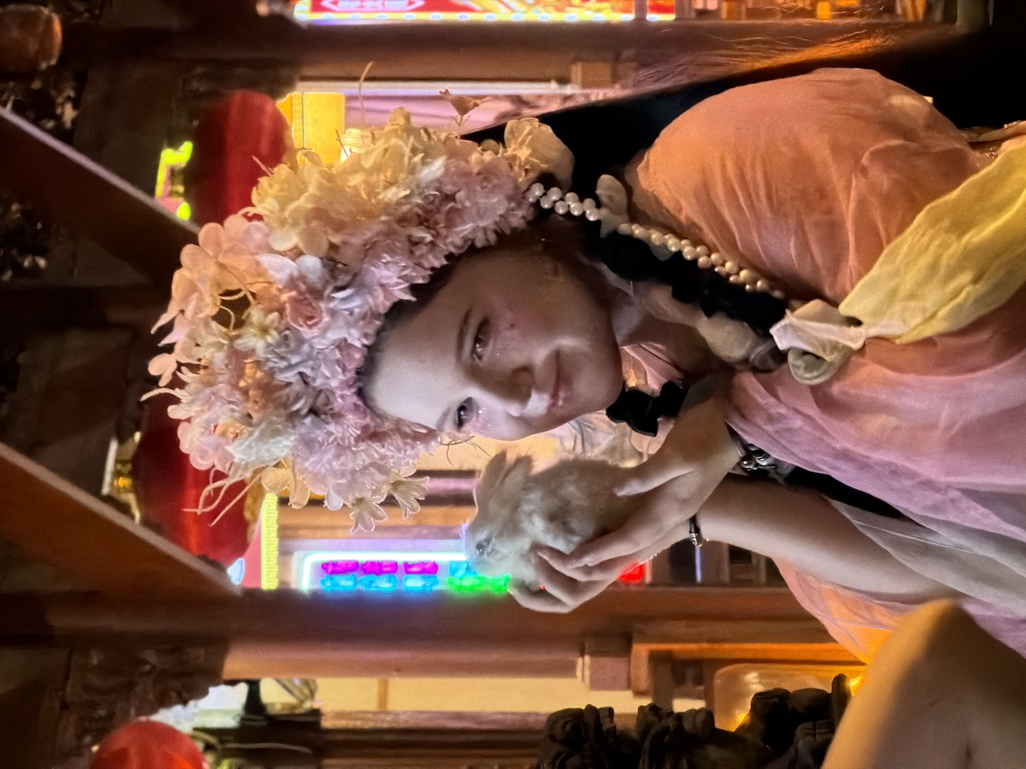 A young Caucasian female with light skin, dark braided hair, and a floral headpiece adorned with pink and white flowers is holding a small, light-colored rabbit in her hands. She is wearing a pink, flowing garment with pearl embellishments hanging from her hair. The background suggests an indoor setting with traditional Chinese architectural elements, including wooden beams and red lanterns. Colorful neon lights are visible in the distance. The overall atmosphere is festive and possibly related to a cultural event. The location is Yuzhong District, China.