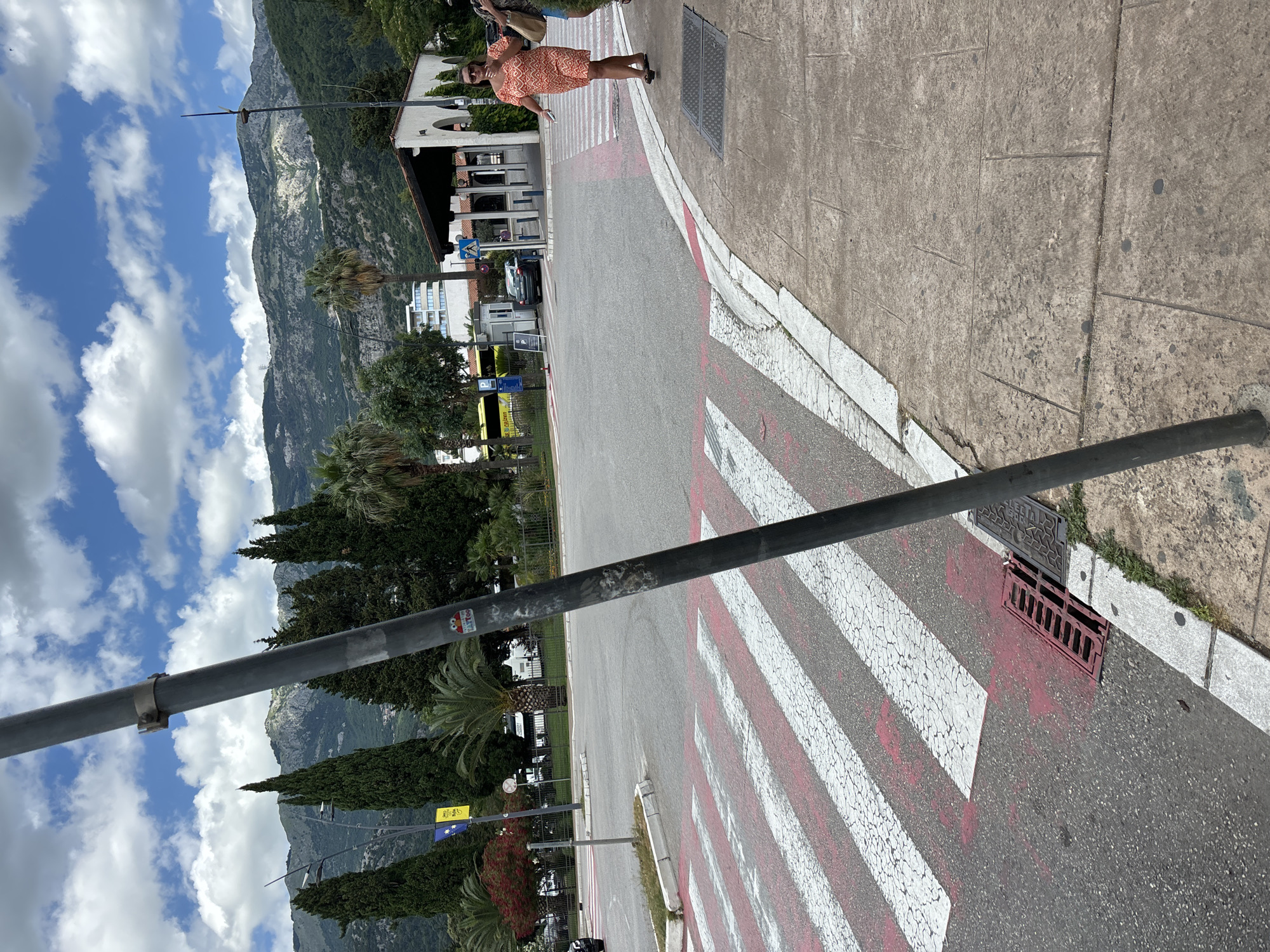 A pedestrian crosswalk with red and white stripes is visible in the foreground, marked with red paint. A dark gray pole stands in front of the crosswalk, obscuring a portion of the image. To the right of the crosswalk is a sidewalk made of stone tiles. Two women are walking on the sidewalk, with one in an orange patterned dress in focus, carrying a brown bag. Behind the women, a building with an arched entrance and a dark roof is visible. In the background, there are trees, a yellow bus, and a range of mountains under a cloudy sky. The scene appears to be in Budva, Montenegro, possibly near a transportation hub or entrance to a facility.