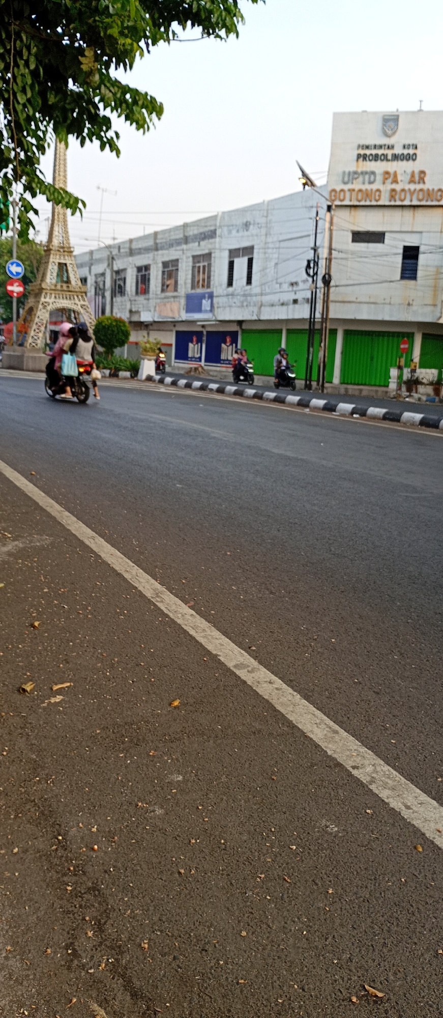 The image depicts a street scene in Probolinggo, Indonesia, featuring a street with a white lane marker. In the background, a building displays signage that reads "PEMERINTAH KOTA PROBOLINGGO UPTD PASAR GOTONG ROYONG." A decorative Eiffel Tower replica stands to the left of the building, and several individuals are seen riding motorcycles. One motorcycle in the foreground carries two passengers, one of whom appears to be wearing a pink headscarf. Another motorcycle with one rider is visible further down the street, as are two more motorcycles with riders approaching from the distance. The street is lined with a pedestrian walkway featuring a black and white patterned border. Green shutters are visible on some of the storefronts. The sky is bright, suggesting daytime.