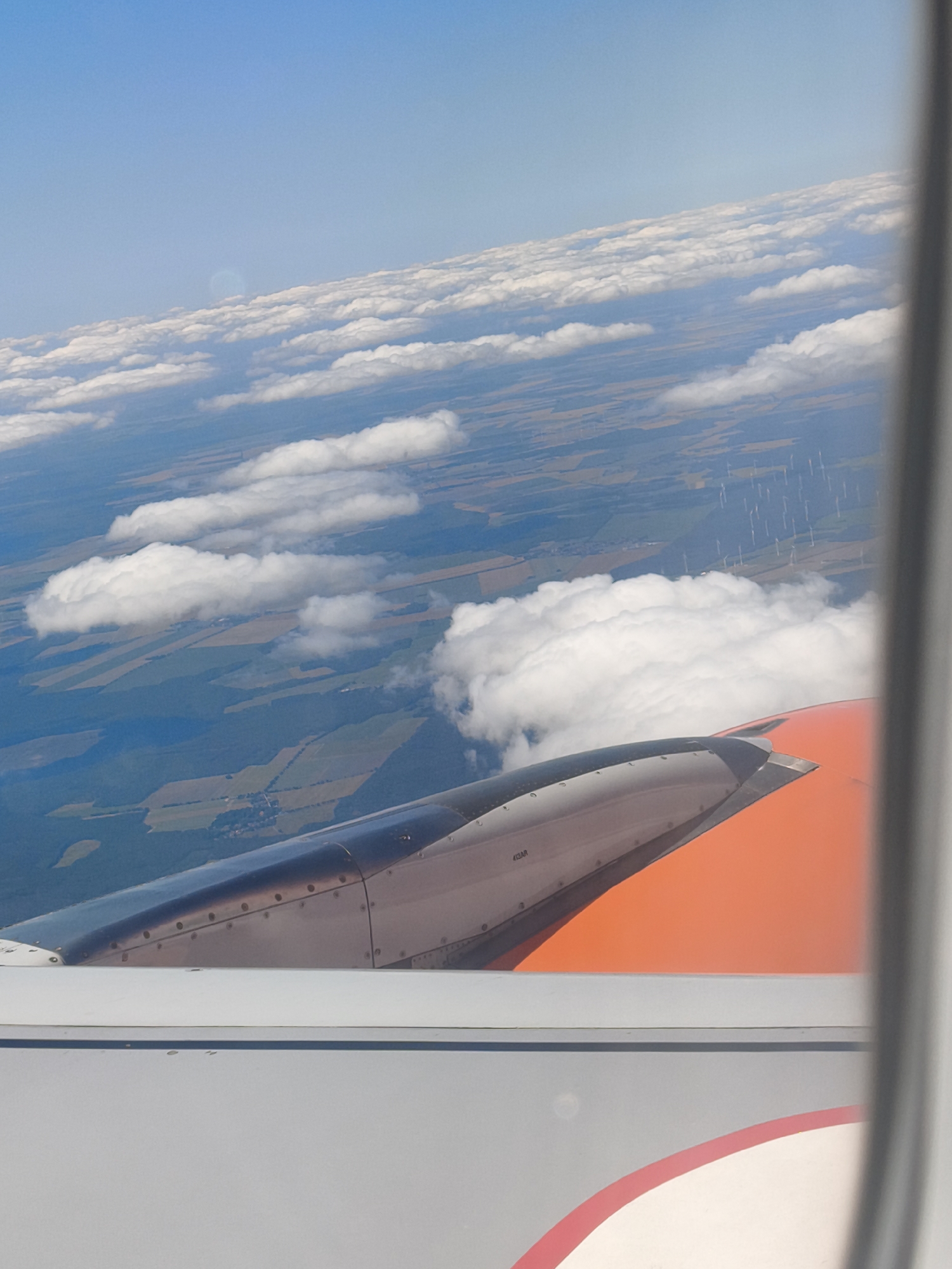 The image, taken from an airplane window, shows a view of the landscape below and the airplane's wing. The landscape features a patchwork of green and brown fields, some with visible patterns suggesting agriculture. There are clusters of trees and buildings, indicative of rural settlements. In the distance, a wind farm with numerous wind turbines is visible. The sky is blue with scattered white clouds, some of which are large and puffy. The airplane's wing is prominently in the foreground, with a metallic section and an orange painted section. The image captures an aerial perspective of the region around Golßen, Germany, a rural area characterized by agricultural land and renewable energy infrastructure.