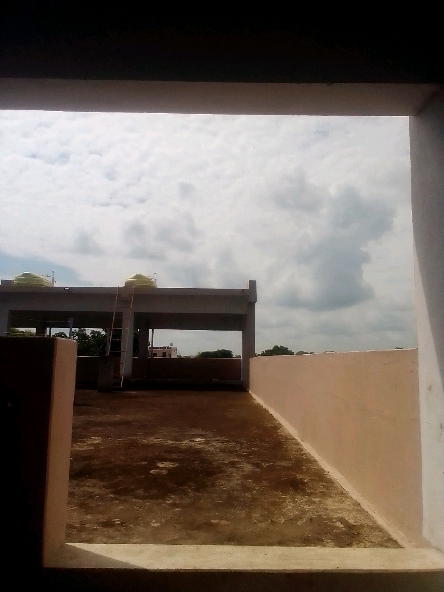 The image depicts a view from a covered balcony or opening, looking out onto a flat rooftop area under a cloudy sky. In the middle ground, there is a concrete structure with two large, light green water tanks on its roof. A ladder leans against the side of this structure. The rooftop itself is paved with a rough, dark material that appears somewhat stained or weathered. To the right, a light-colored wall runs along the edge of the rooftop. In the background, trees and a distant building are visible. The overall scene suggests an urban or semi-urban environment, likely on top of a residential or commercial building. The location is identified as Anjaniya, India.