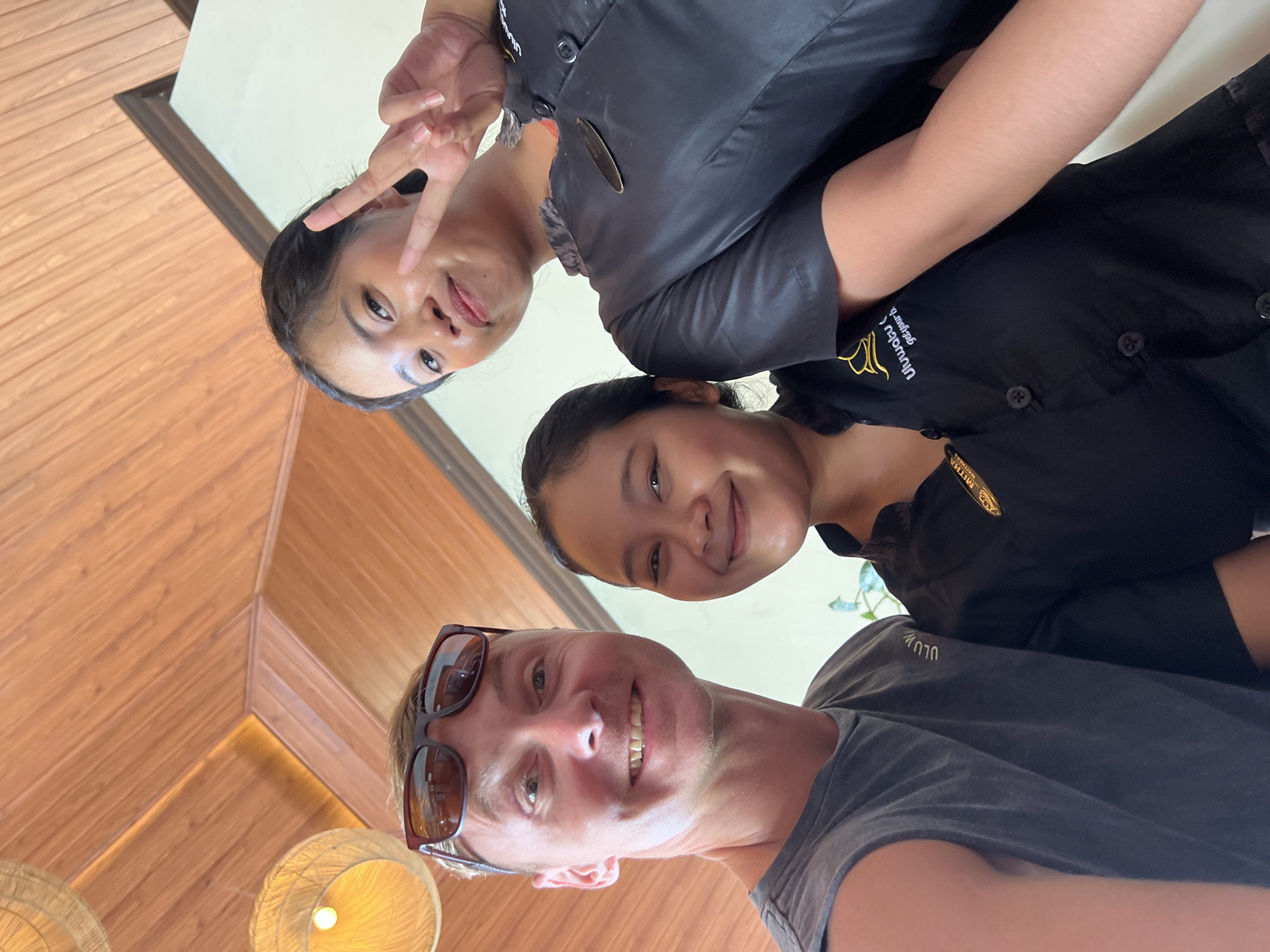 A selfie-style photograph captured indoors depicts a man and two women smiling. The man, situated at the bottom of the frame, wears sunglasses and a gray t-shirt. Positioned above him and to the right is a woman with dark hair pulled back, wearing a black shirt. The woman at the top left of the frame is making a peace sign with her left hand. Both women appear to be employees, possibly waitresses, based on their attire and small embroidered details on their shirts. The interior includes wood paneled walls and hanging woven light fixtures. The photograph was taken in Kuta Selatan, Indonesia, as denoted by the name 'Uluwatu' embroidered on the one woman's shirt.