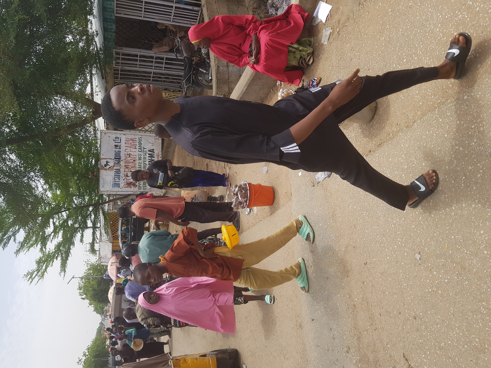 A crowded street scene in Dabawa, Nigeria, shows pedestrians walking and interacting. A young man in black athletic attire and Nike slides stands out in the foreground. Other individuals are visible, some wearing traditional attire, like a woman in a red hijab seated on the ground. Street vendors are present with buckets and items for sale. A faded sign advertises "Dutsinma Trading Co." and references a university campus.