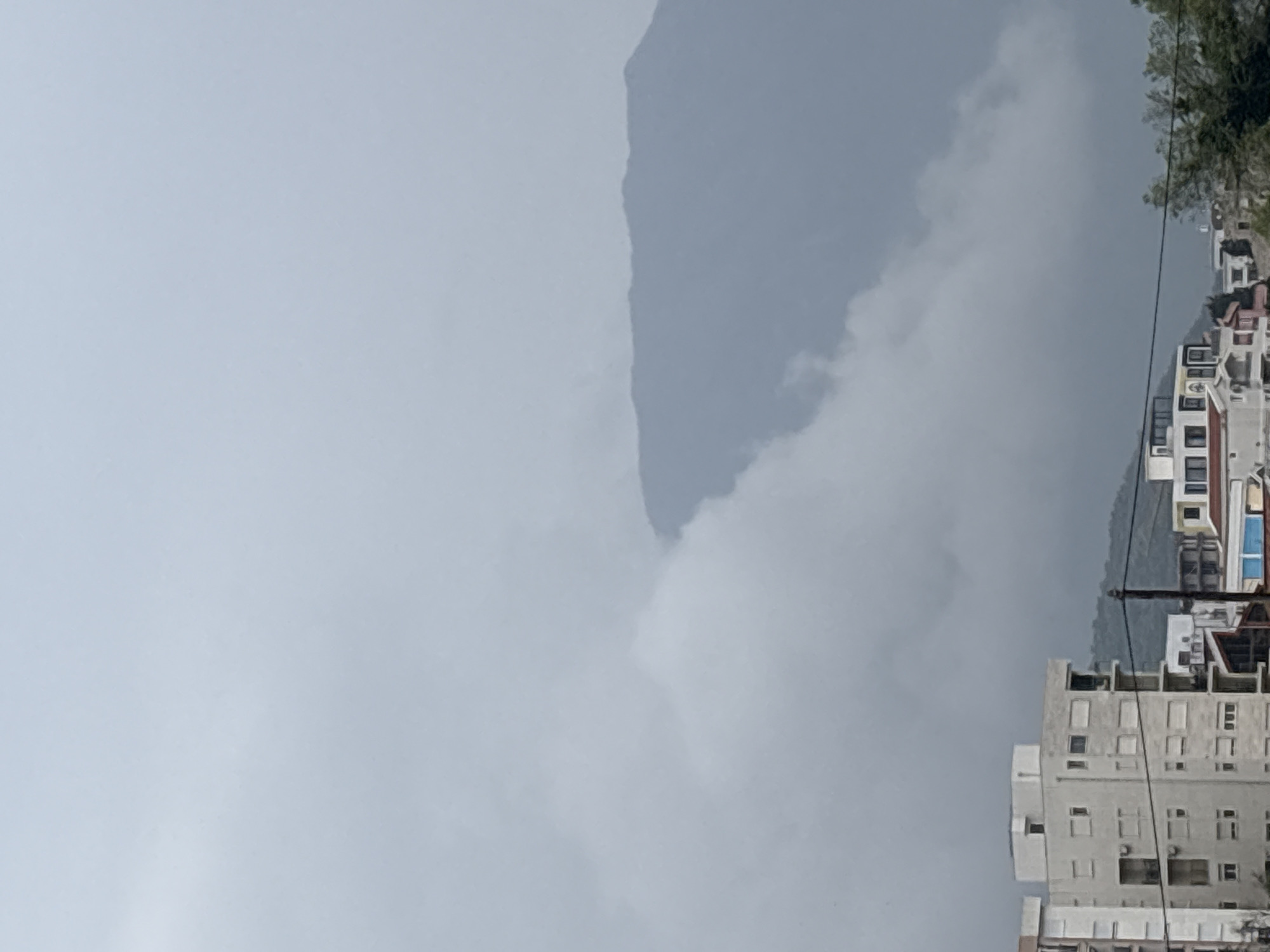 The image, correctly oriented, displays a cloudy day over a town in Bar, Montenegro, with a prominent mountain backdrop. In the foreground, along the bottom and extending up the right side, are several multi-story buildings, likely residential or commercial structures such as apartments or hotels. These buildings feature light-colored facades, numerous windows, and some balconies, reflecting a functional architectural style. Utility cables or power lines run diagonally across the scene, particularly on the right side, in front of the buildings.
The dominant feature of the midground is a large mountain or hill that rises dramatically on the left, its slopes and peak partially veiled by dense clouds or fog. The sky above is entirely overcast, presenting a uniform light grey hue that contributes to the soft, diffused lighting. This extensive cloud cover creates a tranquil yet somewhat muted atmosphere. On the far right, a cluster of green foliage, possibly trees, is visible above some of the buildings, adding a touch of natural color to the otherwise grey and muted palette.
No human activity or specific events are depicted. The scene captures a static landscape of an urban area against a natural mountain setting during an overcast day. There is no visible text in the image.