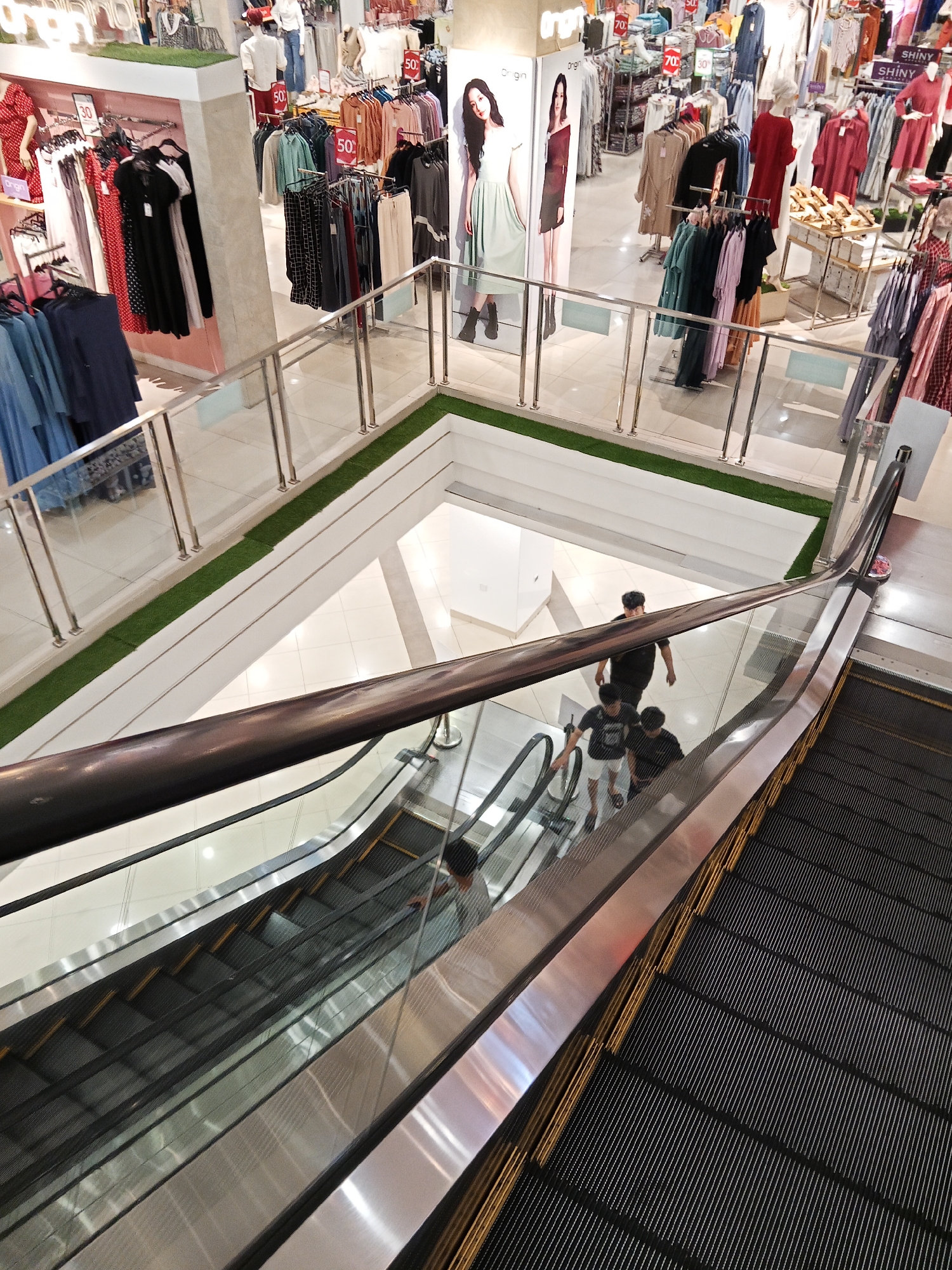 The image depicts an interior view of a multi-level department store, likely located in Lubuklinggau, Indonesia, characterized by its brightly lit aisles and a wide array of clothing displays. In the foreground, an escalator with dark treads and stainless steel railings is visible, with three young men ascending. One man in a black t-shirt leads, followed by two others. Above, a well-lit section of the store showcases racks of colorful apparel, mannequins dressed in various outfits, and promotional signage, some indicating "50% Off." The store appears to be well-stocked, with clothing organized by color and style on hanging racks and shelves. The flooring is a combination of white tiles and a green, grass-like material lining the edges of the upper level. The overall impression is one of a busy retail environment.