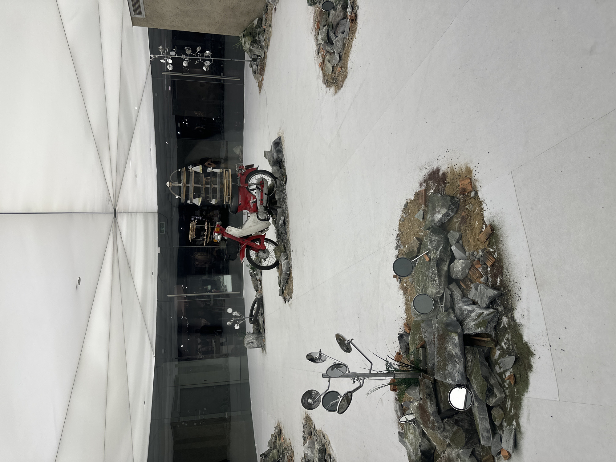 The image shows an indoor exhibit located in Thủ Đức, Vietnam. A red motorcycle with a white seat is parked on a display of rocks and dirt, giving the impression of it being off-road. There are multiple streetlights with circular mirrors standing on similar rocky displays spaced across the area. The background features a modern building with geometric white panels and reflective glass. A shop inside the building is visible through the glass. The exhibit is indoors, suggested by the tiled floor. No people are explicitly visible, although reflections in the glass may show them.