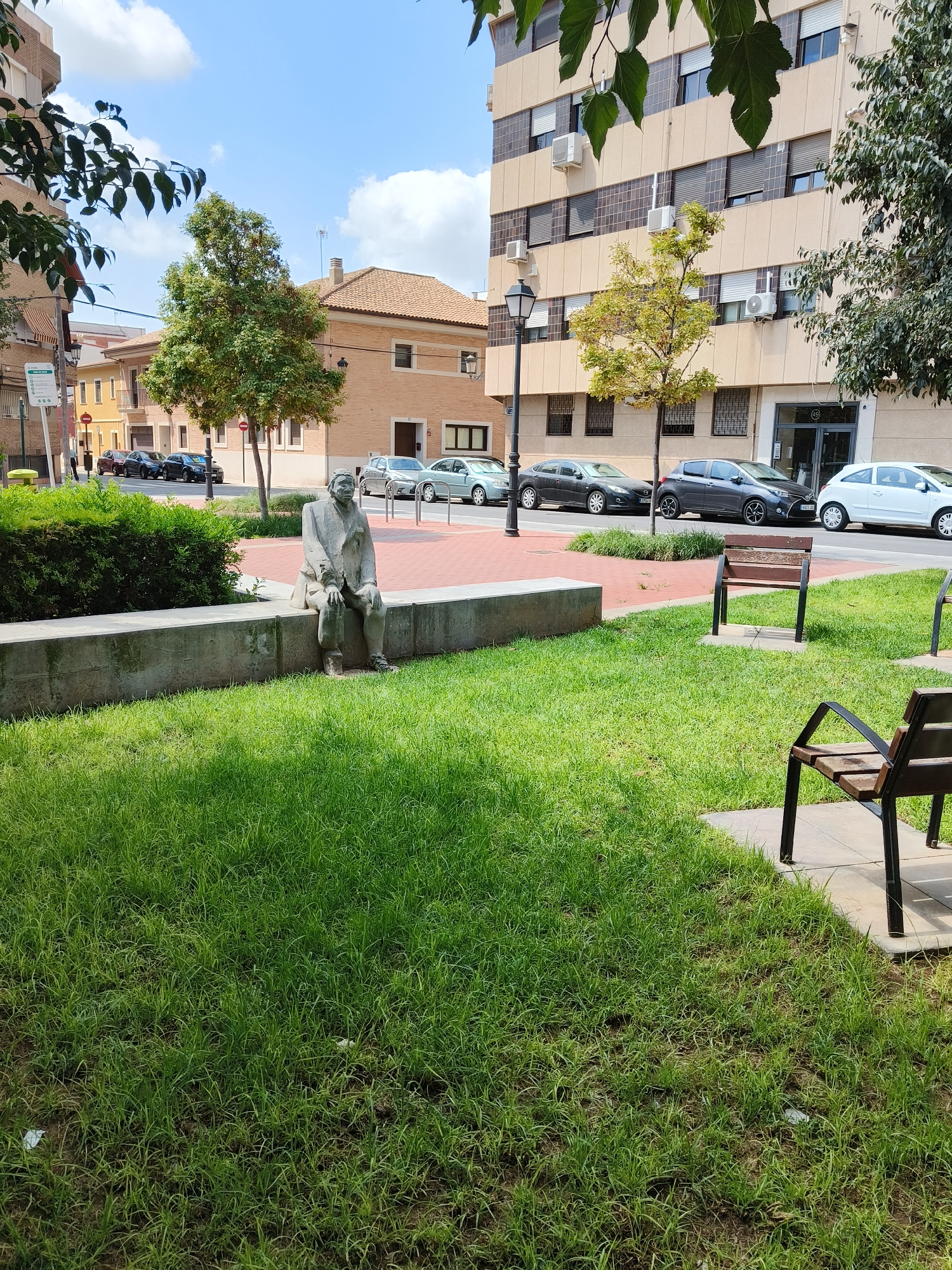 In the city of Benimàmet-beniferri, Spain, a statue of a man is seated on a low concrete wall. The statue, made of a light-colored material, appears to be dressed in a coat and trousers, with a hat on its head. The man is depicted in a relaxed posture, with his hands resting on his lap. The statue is positioned on a grassy area, in front of a thick hedge. Beyond the hedge, a street is visible with several parked cars. A lamppost stands to the left of the statue. In the background, a building with multiple windows and air conditioning units is present, along with other residential buildings. The sky is blue with scattered clouds, suggesting it is daytime. The overall scene depicts a public outdoor space, possibly a park or plaza, with elements of urban infrastructure and greenery.