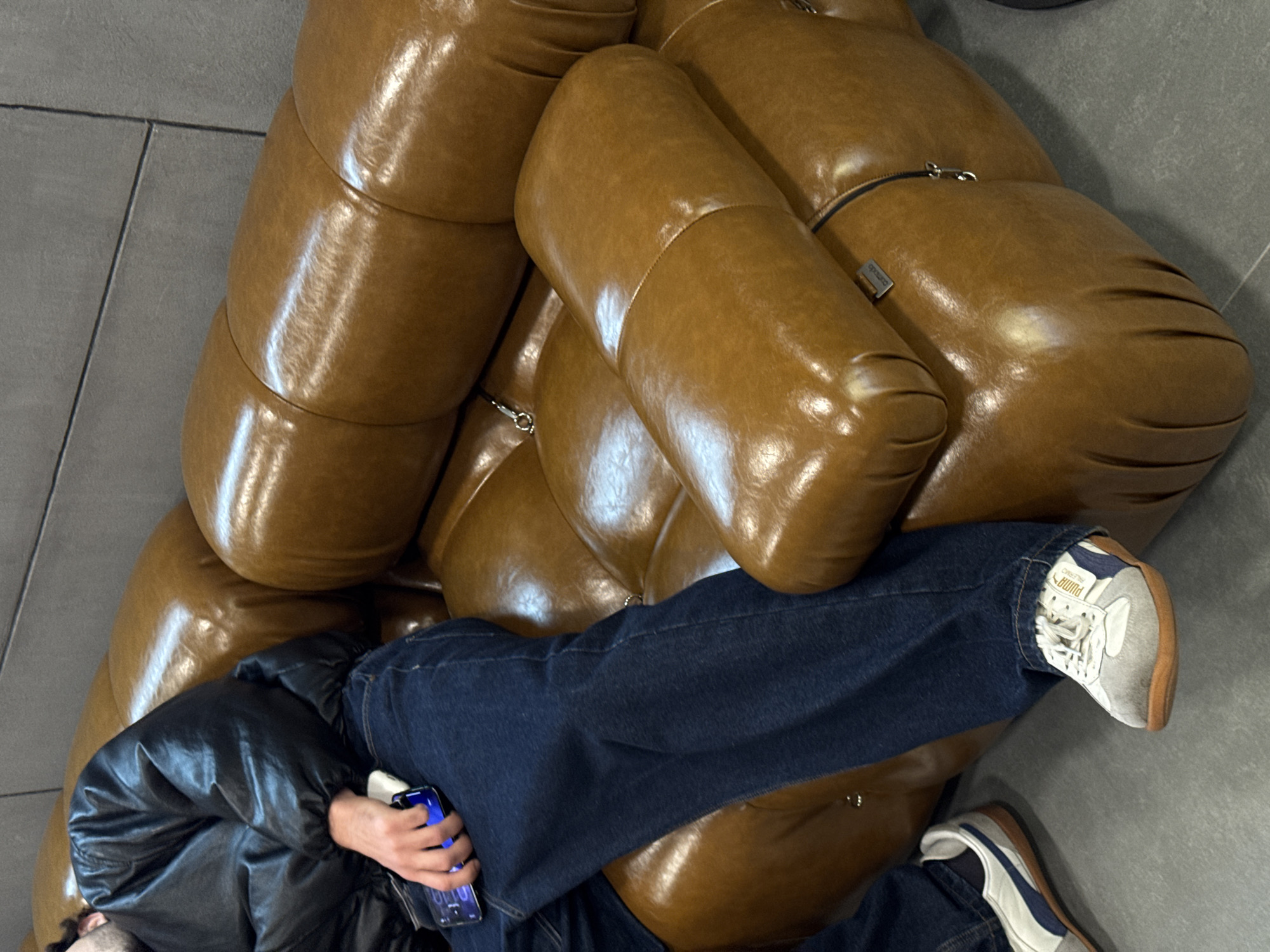 A man in a black leather jacket and jeans reclines on a tan leather puffy couch, appearing to use a smartphone. He is wearing two different Puma sneakers: one is mostly white with a tan sole, and the other is mostly white with a blue stripe and a tan sole. The location is possibly Yerevan, Armenia.