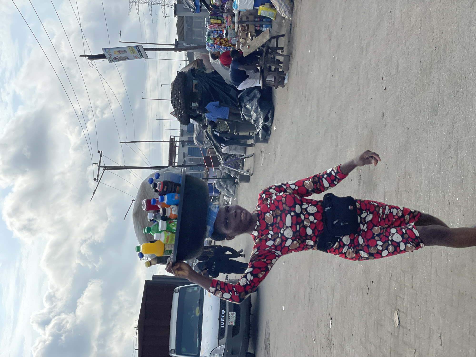 In a bustling market setting in Lekki, Nigeria, a young woman walks purposefully, balancing a large, dark basin filled with various colorful bottles of beverages on her head. She wears a vibrant red dress adorned with a pattern of white, black, and red abstract shapes. A black fanny pack is cinched around her waist. The ground is an unpaved, dusty surface. In the background, a white Iveco van is parked, and other individuals are visible engaged in market activities, with stalls and makeshift structures suggesting a commercial area. The sky above is partly cloudy, with a mix of white and gray clouds. Overhead power lines and utility poles indicate urban infrastructure.
