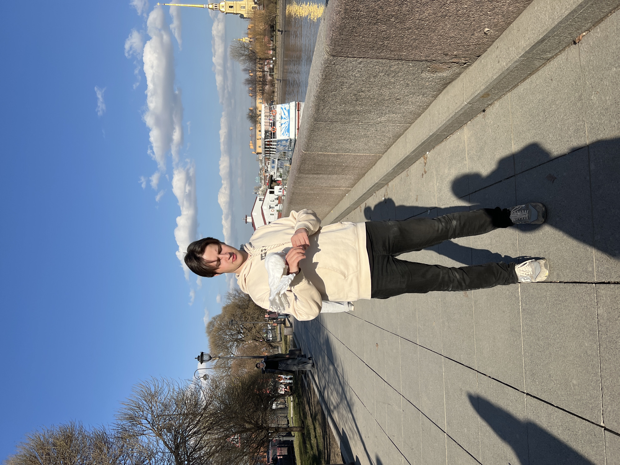 A young male, approximately late teens to early twenties, with dark brown hair, stands centrally on a grey paved pedestrian walkway. He wears a light beige hooded sweatshirt with "Levi's" text on the chest, black jeans, and grey athletic shoes. His gaze is directed downwards at a white pigeon resting on his left forearm, with his right hand positioned near the bird.
To the right of the male, a rough-textured granite embankment wall separates the walkway from the Neva River. Several white and blue passenger boats are visible on the river, with various buildings on the far bank. Prominently in the background, across the river, stands the golden spire of the Saints Peter and Paul Cathedral within the Peter and Paul Fortress, affirming the location as Saint Petersburg, Russia.
To the left of the male, a second paved path runs alongside a treed area with bare branches, indicative of early spring. A street lamp post is visible. A female figure in dark attire walks away from the camera along this path. The sky is bright blue with scattered white cumulus clouds. Long shadows are cast on the paved ground, suggesting a low sun angle.