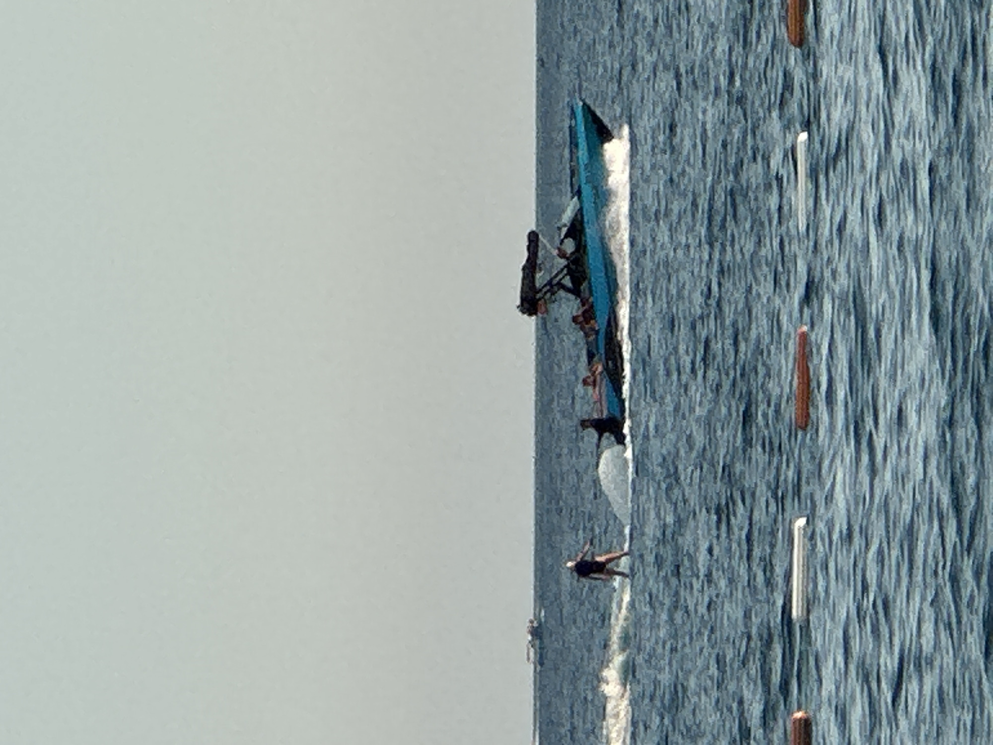 In the city of Boreti, Montenegro, a watercraft activity is depicted on a body of water, likely the Adriatic Sea. The scene is dominated by the expansive sea, with a clear, light-colored sky overhead. In the mid-ground, a bright turquoise boat is moving across the water, creating a white wake. Several individuals are visible on the boat. To the left of the boat, a person is standing on a wakeboard, being pulled by the boat, their body angled as they navigate the water. In the foreground, three buoyant markers, possibly for a water sport course or boundary, are floating on the surface of the water. The overall atmosphere suggests a recreational water sports activity under a bright, overcast sky.