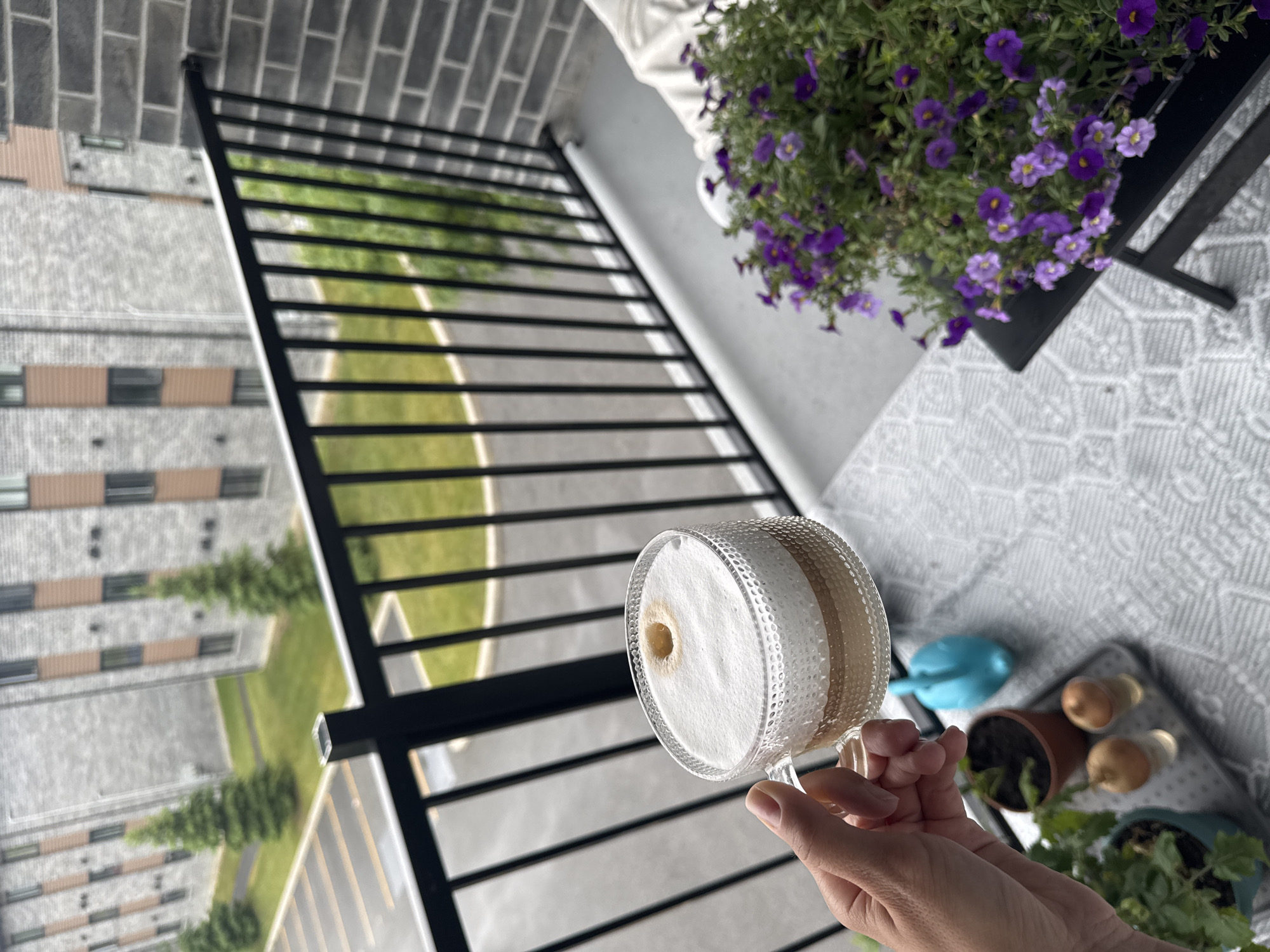 A hand is holding a clear glass mug filled with a frothy beverage, likely coffee. The mug has a textured pattern on its exterior. The beverage has a light tan foam with a small darker circle in the center, suggesting a coffee drink with crema or a swirl of espresso. The hand holding the mug is lightly gripping the handle.
The scene appears to be an outdoor balcony or patio. A black metal railing with horizontal bars stretches across the foreground, creating a layered perspective. Beyond the railing, there is a view of apartment buildings with light-colored facades and dark windows, interspersed with green trees and lawn. The architecture suggests a modern urban or suburban setting.
In the background, on the balcony surface, are several potted plants. One pot holds vibrant purple flowers, possibly petunias or calibrachoa, spilling over the edge. To the right, other small plants and two light brown, round objects, possibly decorative spheres or small potted plants themselves, are visible. A small blue watering can rests on a patterned rug or mat.
The lighting suggests it is daytime, with sunlight casting shadows through the railing onto the balcony floor and the green space outside. The overall atmosphere is calm and relaxed, possibly a moment of enjoying a beverage outdoors. There is no visible text in the image. The location is specified as Gatineau, Canada.