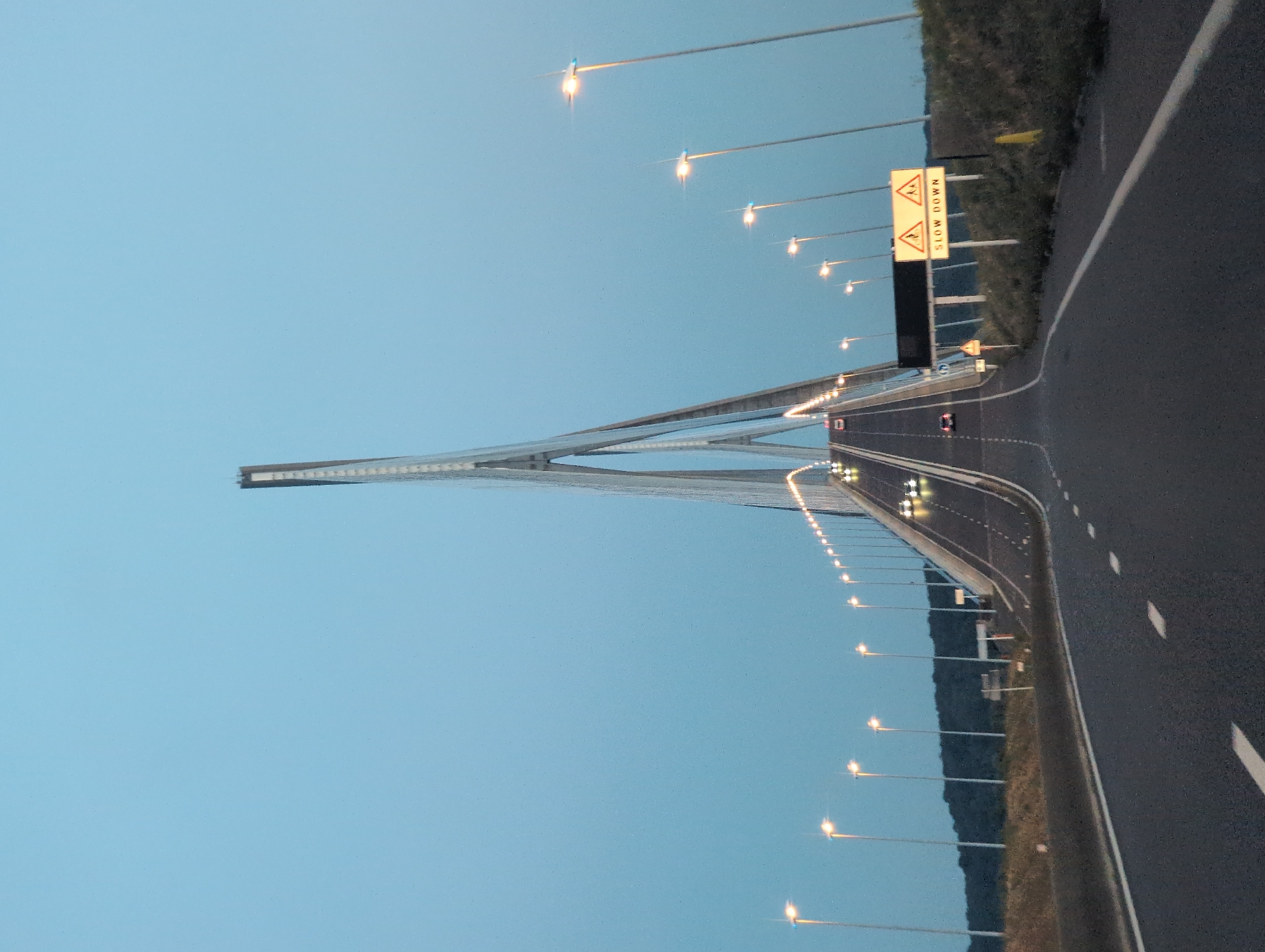 The image shows a view from a vehicle traveling on a multi-lane highway that leads onto a large cable-stayed bridge. The bridge has a prominent central pylon with cables supporting the roadway. The sky is a clear, pale blue, suggesting dusk or dawn. Streetlights are illuminated along the sides of the highway and bridge, casting a warm glow. Several vehicles are visible on the road, some ahead and some approaching. On the right side of the road, there are traffic signs. One sign displays two yellow triangles with figures of people walking, and the text "SLOW DOWN" below. The surrounding landscape on the left appears to be a grassy embankment with trees in the distance. The location is identified as the Pont de Normandie in Sandouville, France.