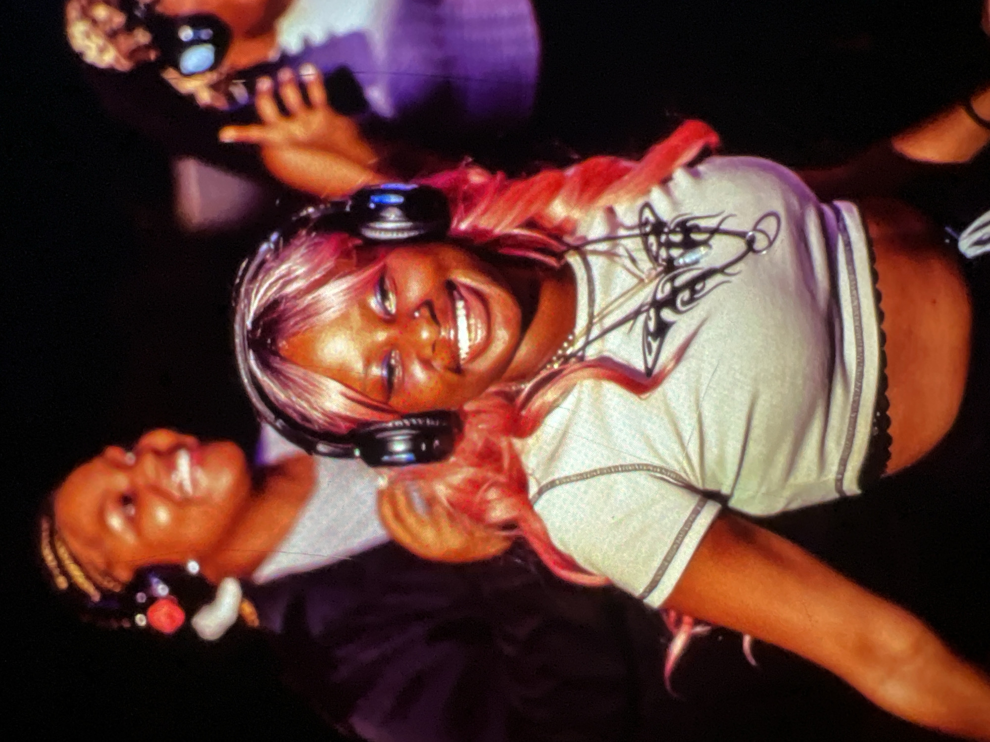 The image captures a medium shot of two Black women wearing headphones in what appears to be a dimly lit indoor setting, possibly a club or party. The woman in the foreground, with bright pink hair, smiles widely at the camera. She is wearing a light-colored cropped t-shirt with a black graphic design on the front and a black lace trim at the hem. She has a nose ring and is wearing a necklace. Behind her and to her left, another woman with braided hair is also wearing headphones and appears to be laughing. She is wearing a dark jacket over a light-colored top. The background is dark, with some blurred purple and white lights, suggesting a vibrant and energetic atmosphere. There are no visible Lagos landmarks or specific environmental cues to pinpoint the location within Lagos, Nigeria.