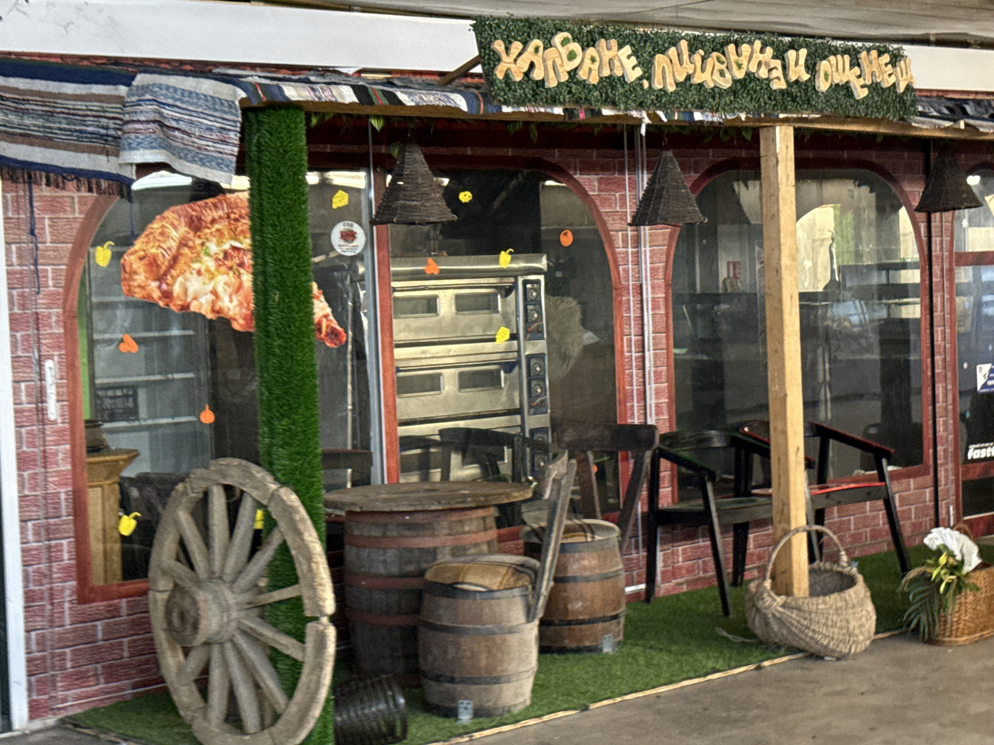 A low-angle, medium shot captures an outdoor food stall with a rustic, somewhat whimsical aesthetic. The stall's facade is constructed from red brick-patterned material, with large arch-shaped windows that offer glimpses of the interior, which appears to house bakery ovens and shelves. Above the windows, a banner with wooden, carved lettering in a foreign language is affixed, partially obscured by a green leafy garland. Decorative elements include hanging cone-shaped lamps, a large wooden wagon wheel leaning against the stall, and two barrels repurposed as seating with cushions. A tall, artificial green plant stands to the left of the scene. The ground is covered in artificial turf, with a woven basket and a floral arrangement placed near the foreground. A large, slightly cartoonish image of a pizza slice is displayed prominently on the left side of the facade, suggesting the type of food offered. The overall impression is that of a charming, perhaps family-run, establishment aiming for a unique visual identity.