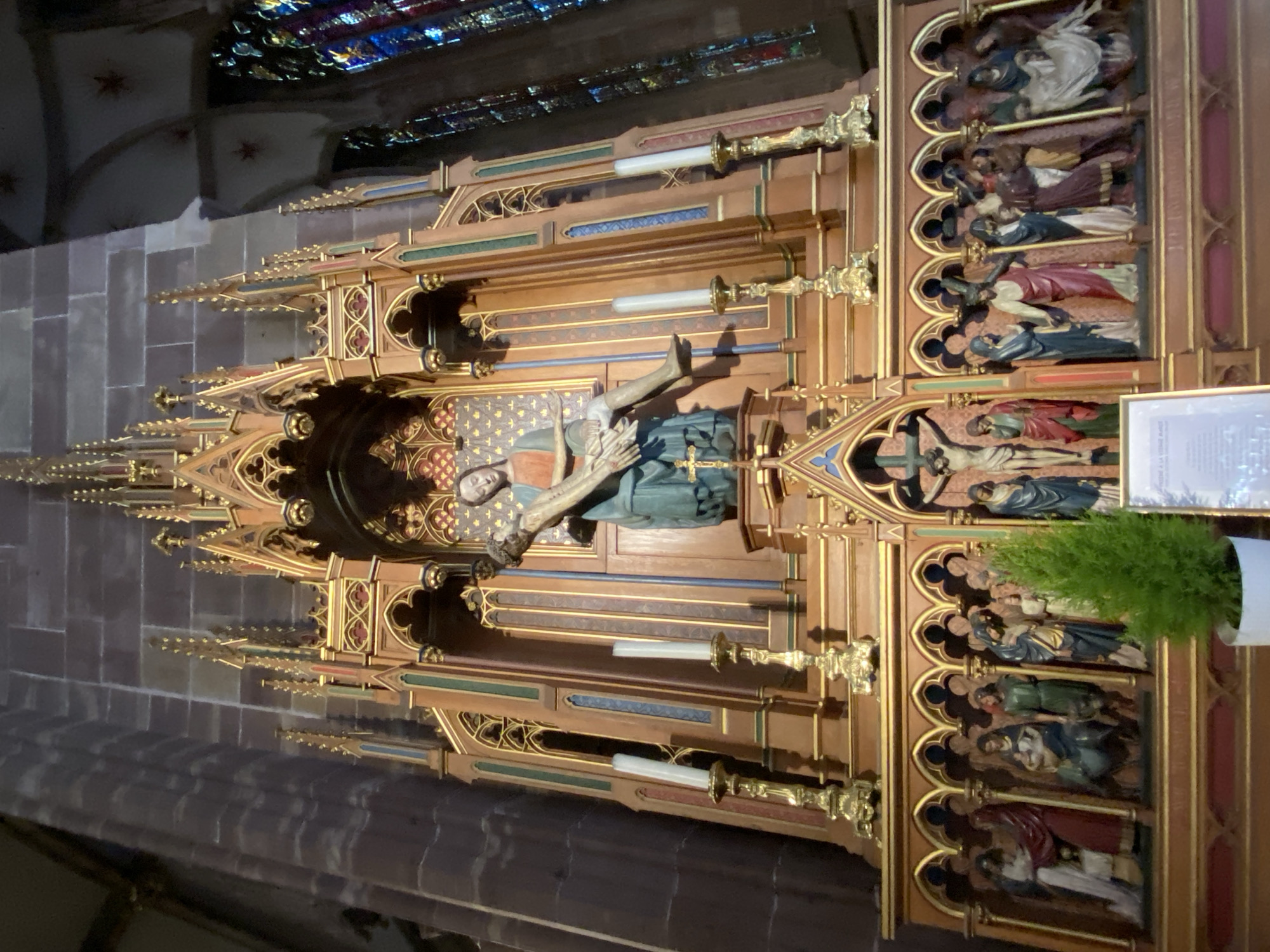 The image displays a large, intricately carved wooden altarpiece or reredos, situated within a church or cathedral in Strasbourg, France. The piece is a rich example of Gothic artistry, featuring multiple religious sculptures and ornate architectural details.
At the center of the altarpiece, a prominent carving depicts a recumbent figure, likely the dead Christ, being mourned or supported by a sorrowful female figure, possibly the Virgin Mary. This central scene is flanked by elaborate architectural elements, including spires, finials, and pointed arches, all adorned with gold leaf and painted accents in blue, red, and green.
To the right of the central tableau, and extending downwards, are several rows of smaller sculpted scenes. Each scene is housed within its own arched niche and contains multiple figures, depicting various events, most likely from the Passion of Christ or other biblical narratives. One visible scene clearly shows a figure on a cross.
Decorative motifs, including fleur-de-lis and geometric patterns, enhance the wooden structure. Two white electric candles in decorative brass holders are integrated into the altarpiece. A small green potted plant rests on the far right side, adding a contemporary touch.
In the background, vaulted stone ceilings and sections of vibrant stained-glass windows are visible, indicating the grandeur of the sacred space. The interior lighting appears dim, contributing to a solemn and reverent atmosphere. A small, white rectangular plaque with text is partially visible on the right, but the writing is too blurry to be read.