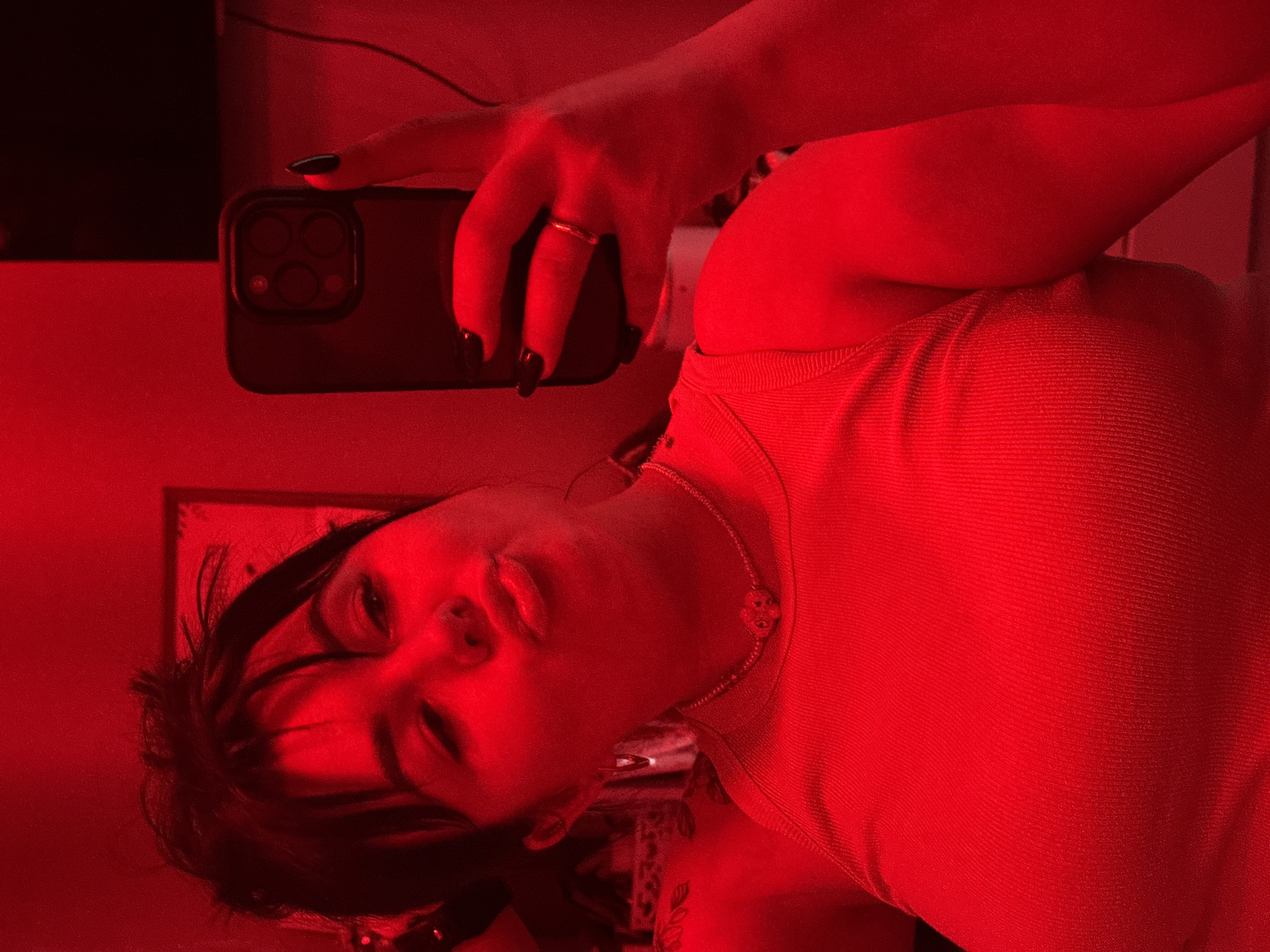 A person is taking a selfie with a smartphone. The subject has black nail polish and wears a ring on their index finger. The environment is lit with red light, casting a uniform hue across the scene, which impacts visibility and detail. The specific location of Chaikovsky, Russia, cannot be determined from the visual information.