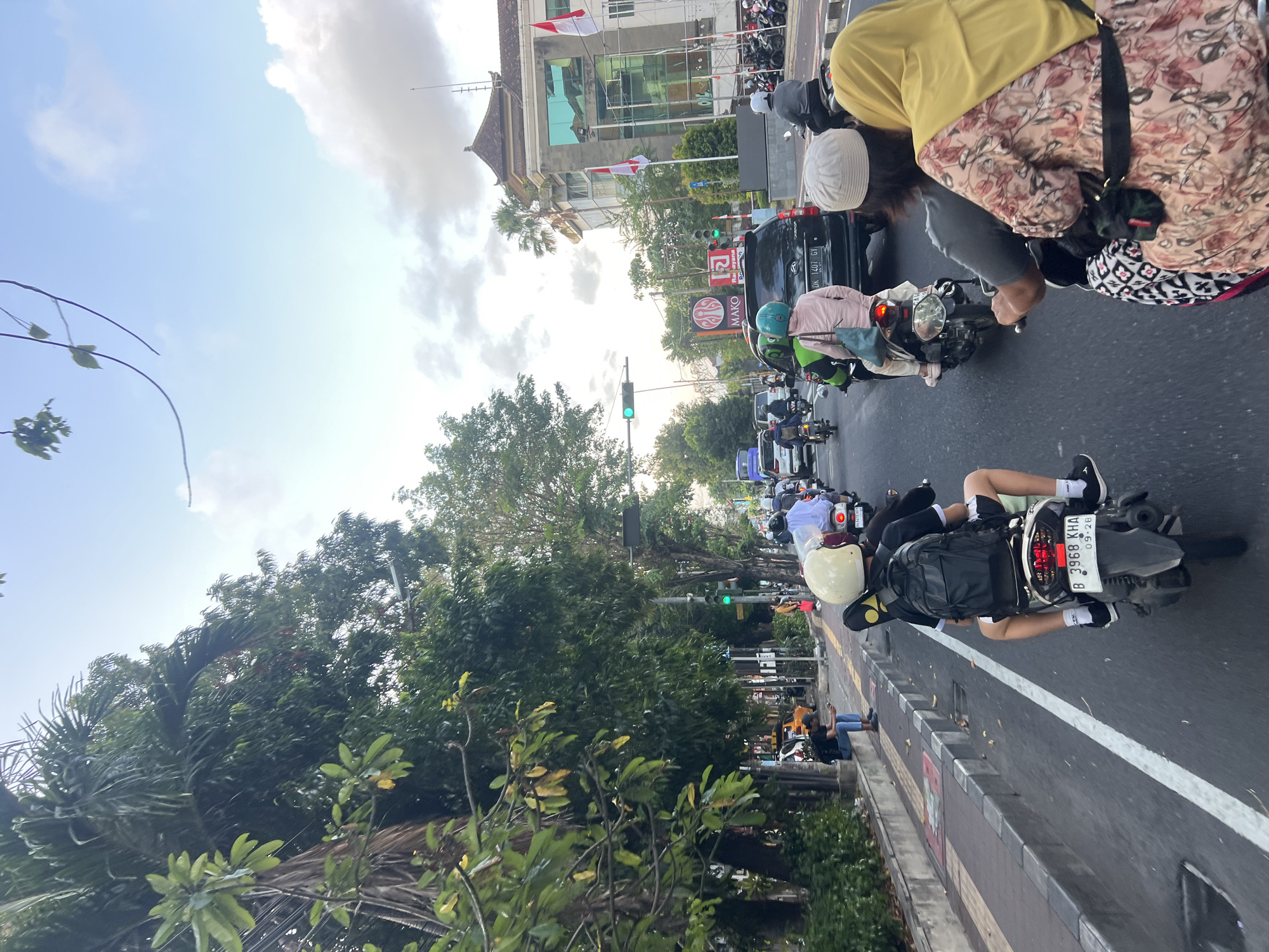 A group of people are riding motorcycles in a traffic jam on a street in Denpasar, Indonesia. The image is taken from the perspective of someone on the back of a motorcycle, looking forward. The sky is overcast with a few patches of blue. Trees line the street on the left, and a building with a traditional Balinese roof is visible on the right. There are several motorcycles and cars on the road. One person on a motorcycle is wearing a yellow helmet and a black backpack. Another person is wearing a pink shirt and a blue helmet. Two people are riding on the back of a motorcycle, with the person in front wearing a grey shirt and a white cap, and the person in the back wearing a yellow hijab and a patterned dress. Traffic lights are visible, with one appearing to be green. A yellow taxi is parked on the side of the road. A person is sitting on a curb on the sidewalk, looking at their phone. Indonesian flags are hanging from the building on the right.