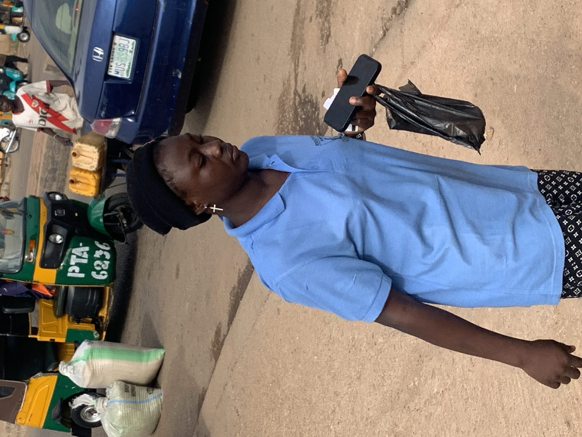 The image depicts a street scene in Gura Top, Nigeria. In the foreground, a young Black woman wearing a light blue polo shirt and a black beanie is walking while holding a smartphone in her right hand and a black plastic bag in her left hand. She has a cross earring in her left ear. Behind her, a blue sedan with a Nigerian license plate is parked. To the left of the woman, a three-wheeled vehicle, commonly known as an "okada" or tuk-tuk, is visible, with two large sacks of goods placed next to it. In the background, a man in a white and red jersey stands near a cart with two yellow jerrycans, and other vehicles, including scooters and more three-wheeled vehicles, are present on the street. The ground is paved with rough asphalt, and the overall environment appears to be a busy marketplace or roadside commercial area.