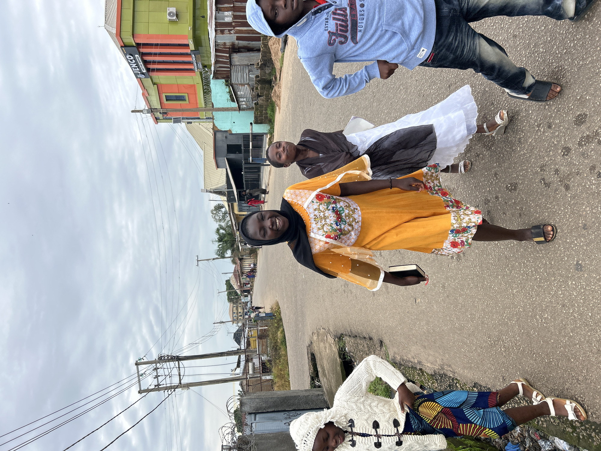 Four young people are walking on an asphalt road in Jos, Nigeria. The person on the far left is wearing a white knit sweater with a hood and a colorful patterned skirt. The person next to them, on the left, is wearing a bright orange dress with sheer sleeves and a black headscarf. They are smiling and holding a small black book. The person in the middle is wearing a brown and white outfit and appears to be looking ahead. The person on the far right is wearing a light blue hooded sweatshirt with "Full House Motors" written on it and dark jeans. They are also looking forward. In the background, there are buildings, a street light, and power lines. The sky is overcast.