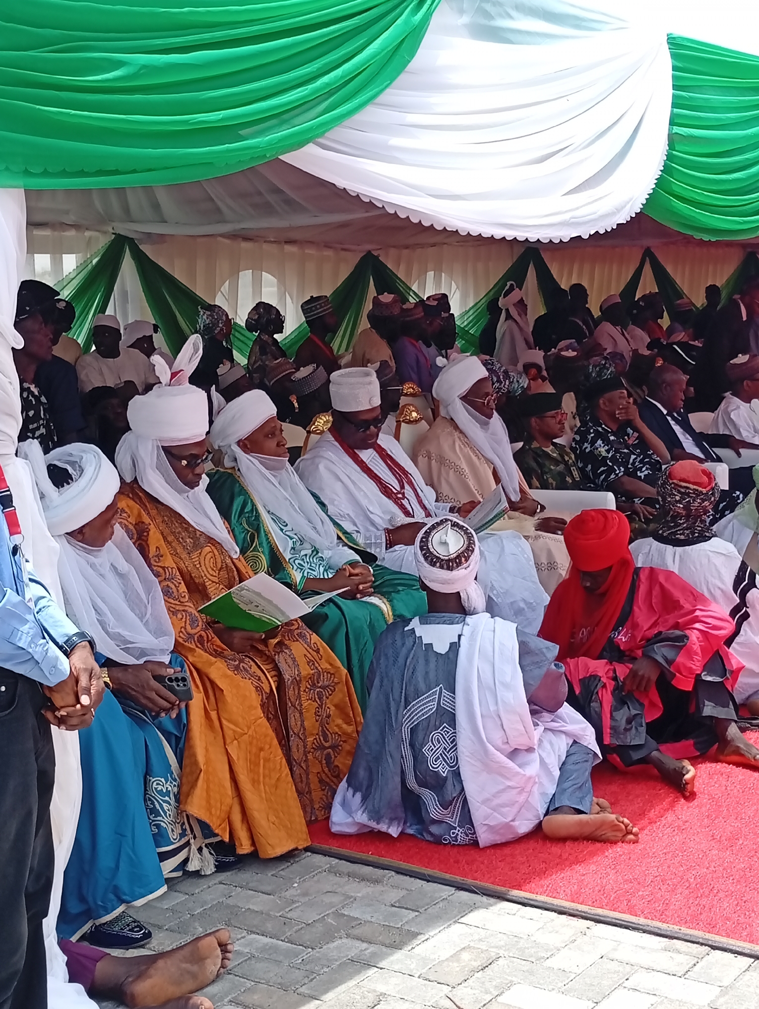 This image depicts a formal outdoor gathering under a large tent, vibrantly decorated with white and green draped fabric overhead and along the sides, evocative of Nigeria's national colors. The event takes place in Unguwan Sanusi, Nigeria, likely during the daytime under bright conditions, with the tent providing shade.
The scene is populated by numerous men, primarily adults and elders, dressed in a variety of traditional Northern Nigerian attire. Many wear elaborate flowing robes (kaftans or boubous) in colors such as orange, gold, green, and white, complemented by white turbans or distinctive caps. Several key figures are seated in rows on white and gold-accented chairs. In the foreground, two younger individuals are seated on a red carpet: one in a striking red ensemble and another in a grey and white patterned garment with a white head wrap. To the far left, a person is partially visible in a blue shirt and dark trousers, holding a mobile phone. Further back, individuals in various attire including a camouflage uniform are seated.
Attendees appear engaged and attentive. Several are observed reading from open books or leaflets, one with a green cover, suggesting a focus on speeches, religious texts, or program materials. The overall atmosphere appears respectful and solemn, indicative of a significant public, traditional, or religious ceremony. The ground under the tent is covered by a red carpet, while outside, grey interlocking pavement is visible. There is no legible text visible on any of the books or other objects.