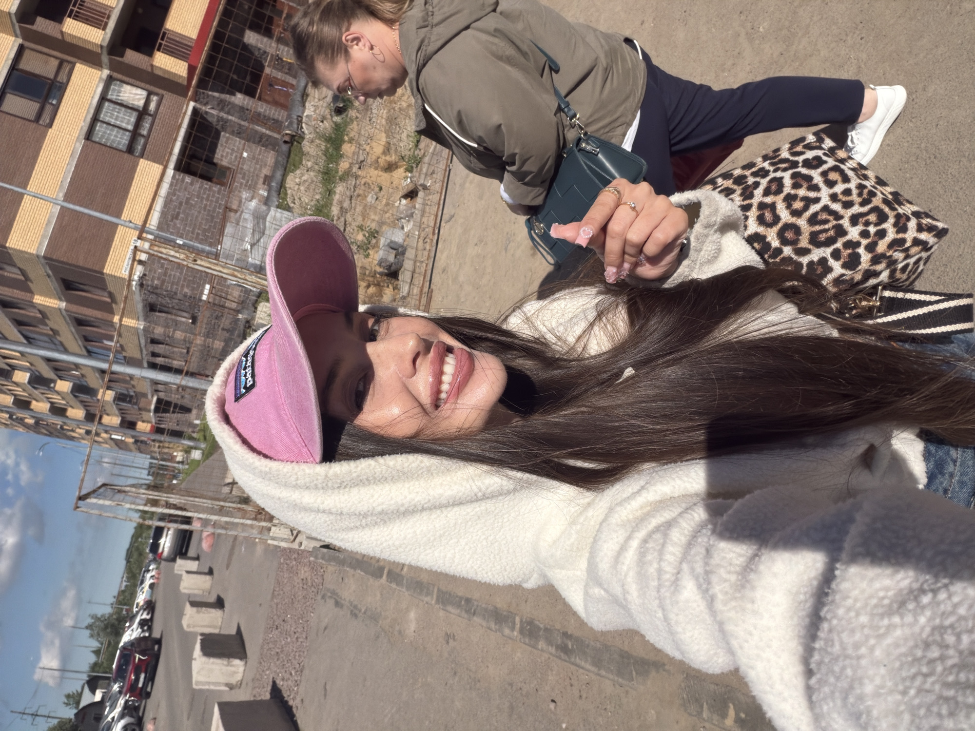 A young woman wearing a white hooded jacket and a pink baseball cap takes a selfie outdoors in a city environment. She has long dark hair, is smiling and sticking her tongue out. In the background, another woman in a green jacket and glasses is visible. The setting appears to be a construction site with a partially built apartment building. Cars are parked in the background, and concrete barriers are along the sidewalk. The lighting suggests it is daytime with a sunny sky. The location is Saint Petersburg, Russia.
