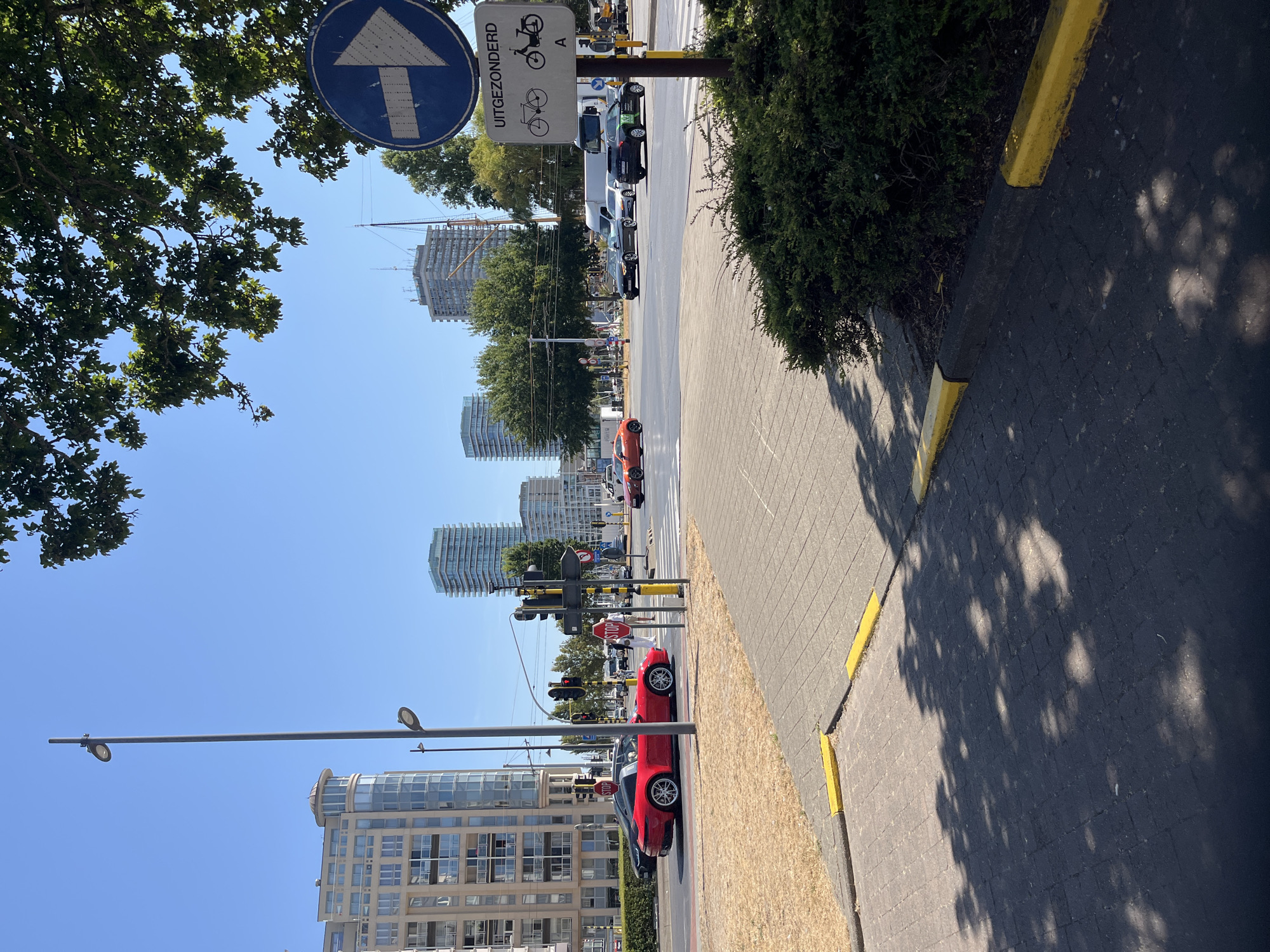The image captures a street scene in Ostend, Belgium, on a clear, sunny day. In the foreground, a red convertible sports car is stopped at a traffic light next to a pedestrian crossing. The car is positioned on the right side of the road, which appears to be a multi-lane intersection. Beyond the intersection, several tall buildings, characteristic of a city skyline, are visible, including modern apartment complexes and a building with scaffolding. Trees line the street, and a blue traffic sign with a white arrow indicating a right turn is prominently displayed. Another sign below it shows a bicycle and a moped with the word "UITGEZONDERD," meaning "exempted" in Dutch. The pavement is made of interlocking bricks, with yellow painted curbs. The overall environment suggests a typical urban setting with traffic and pedestrian activity.