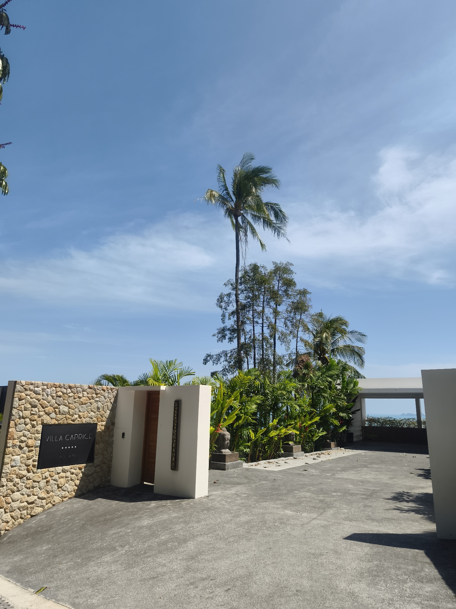 The image shows the entrance to "VILLA CAPRICE" in Ko Samui District, Thailand. The entrance features a stone wall on the left with a black sign displaying the villa name and five stars. To the right of the stone wall is a white structure with a wooden door and decorative elements. Beyond the entrance, lush green tropical vegetation, including palm trees, is visible. In the background, an open-air structure frames a view of the ocean and distant islands under a clear blue sky with scattered clouds. The ground in front of the entrance is paved with grey concrete. Two statues, one appearing to be a Buddha head, are positioned amongst the foliage to the right of the entrance.