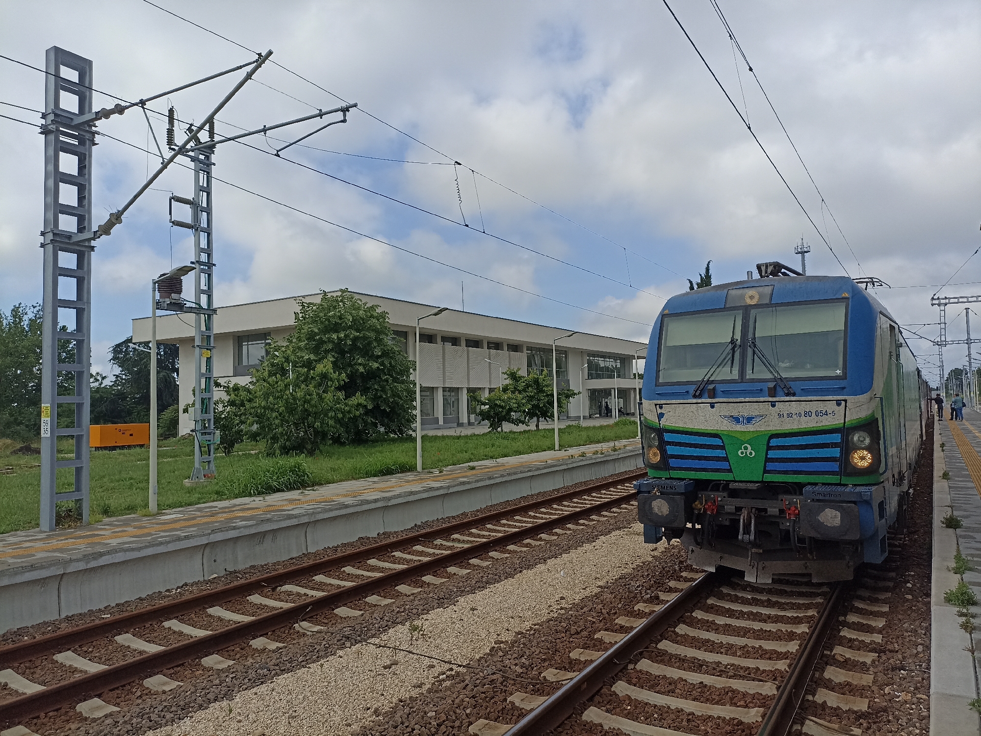 This media file captures a daytime scene at a railway station, likely in Chirpan, Bulgaria, as indicated by the context. The image is dominated by a modern electric locomotive, positioned on the right track, facing the viewer. The train is primarily blue, green, and white, featuring a distinctive winged logo and interlocking circles symbol on its front. Visible text on the front of the locomotive includes the identification number "91 52 10 80 054-5" and the brand names "SIEMENS" and "Smartron." Its headlights are illuminated.
To the left of the train, a set of railway tracks runs parallel to a concrete platform. The tracks consist of metal rails, wooden or concrete sleepers, and gravel ballast. The platform features yellow tactile paving strips. Beyond the platform and tracks, a modern, light-colored building, possibly the station building, stands amidst green grass and trees. The building has large windows and a somewhat minimalist architectural style.
The sky above is mostly overcast with scattered white clouds, suggesting a cloudy but bright day. Overhead, a complex network of electrical lines (catenary system) is supported by tall, metallic lattice-work poles. One pole on the left has a small yellow plate with the numbers "59 / 35" vertically arranged. Another pole nearer the center has various insulators and electrical equipment. In the background, behind the left platform and trees, a yellow rectangular object, possibly a generator or container, is visible. On the far platform, to the right of the train, a few indistinct figures (people) can be seen standing. The train appears to be stationary, possibly at a stop.
