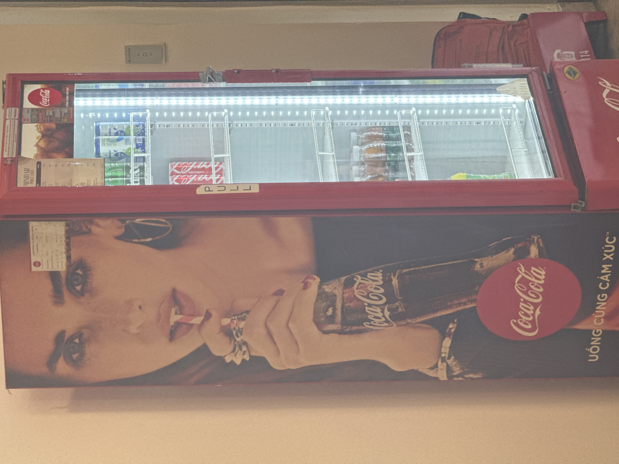 The image shows a Coca-Cola branded vending machine located in a commercial establishment in Hanoi, Vietnam. The machine is primarily red and features a large advertisement on its left side depicting a woman drinking a Coca-Cola. The vending machine itself is lit from within, displaying various beverages on its shelves, including cans of Coca-Cola and other drinks. A "PULL" sign is visible on the front of the machine. The overall environment appears to be indoors, with a plain wall as the background. The lighting is somewhat dim, with the internal lights of the vending machine being the brightest source of illumination.