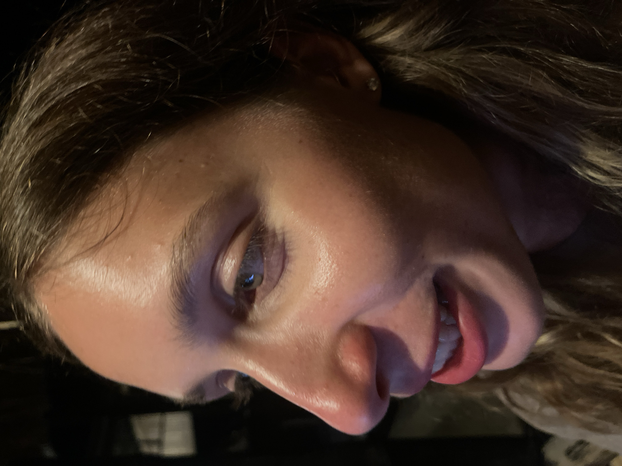 The image is a close-up, side profile of a young woman's face, taken in Kertasari, Indonesia. Her brown hair is partially visible, framing her face, which is illuminated by warm light that highlights the sheen of her skin. She is wearing light-colored lipstick and her mouth is slightly open, revealing her teeth in a subtle smile. Her right eye is visible, with a green iris. She has a small diamond stud earring in her right ear. The background is dark and indistinct, suggesting an indoor setting with low lighting conditions.