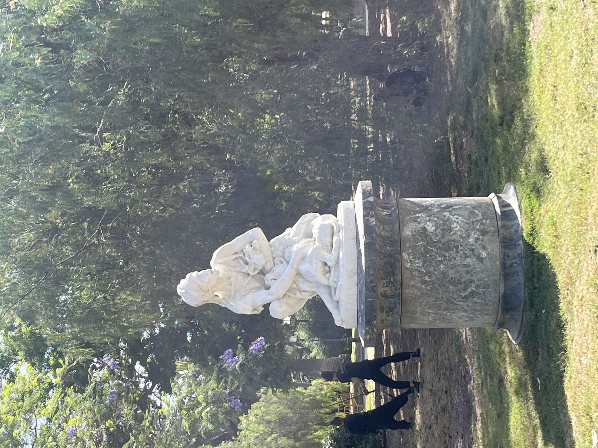 In a sun-dappled park, a white marble statue of a crouching figure holding a small animal stands on a stone pedestal. The figure appears to be female, with a band wrapped around her head. Behind the statue, two individuals dressed in black, one with a sword, are engaged in what appears to be a practice session. The park features lush green grass, trees with dense foliage, and some purple flowers visible. The overall scene suggests a public park setting, possibly a historical garden or plaza. This image was taken in Parc de la Ciutadella in Barcelona, Spain.