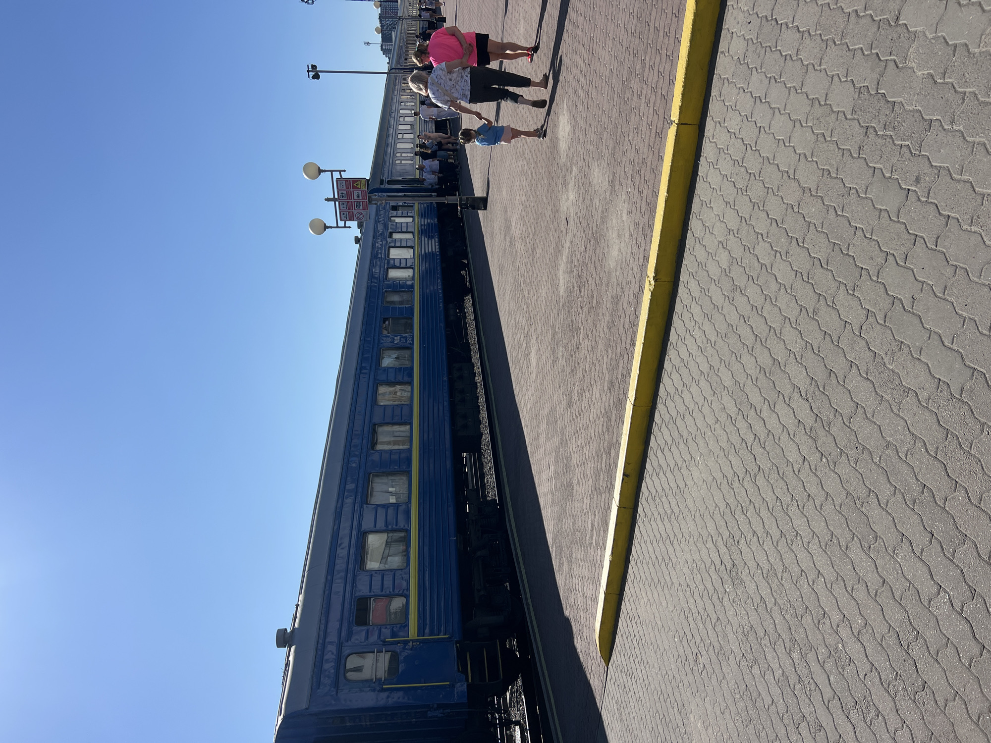 This is a street-level, full-frame shot of a blue train at a station platform on a clear, sunny day. The train is the primary subject, stretching across the frame from left to right. It is a passenger train, with multiple windows visible along its side, some with curtains. A bright yellow stripe runs horizontally along the train's body. The platform is paved with interlocking concrete bricks and has a distinct yellow curb along the edge.
In the foreground, a yellow curb is prominently featured, separating the platform from the lower section of the image which is also paved with interlocking bricks. In the mid-ground, several people are visible on the platform, near the train. Two women and a young child are walking away from the camera towards the train. The child is holding the hand of one of the women. Further down the platform, near the train entrance, a group of people are standing, appearing to be waiting or boarding.
In the background, several lampposts with globe-shaped lights are visible, as well as a sign with various pictograms. The sky is a clear, bright blue, indicating a cloudless day. The scene appears to be a train station in Ivano-Frankivsk, Ukraine, given the architecture and the presence of a train.