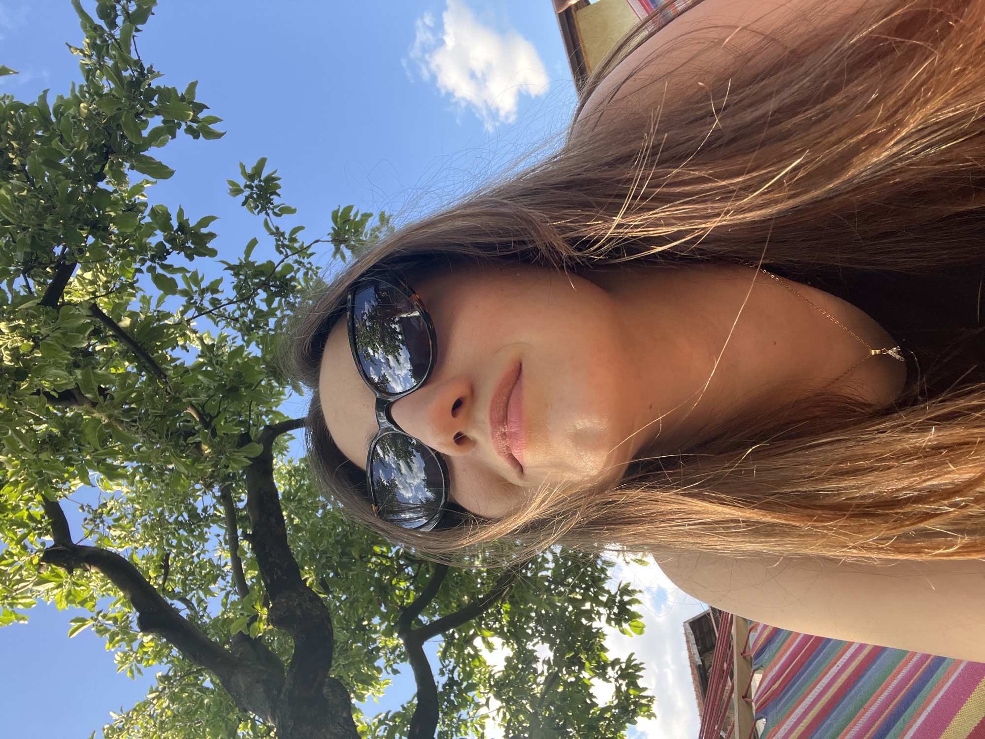 A woman with long brown hair and sunglasses is in Busmantsi, Bulgaria. The sunglasses reflect a blue sky with clouds. She wears a gold necklace. A tree with green leaves is to her left. A building with a colorful striped awning is on her right.