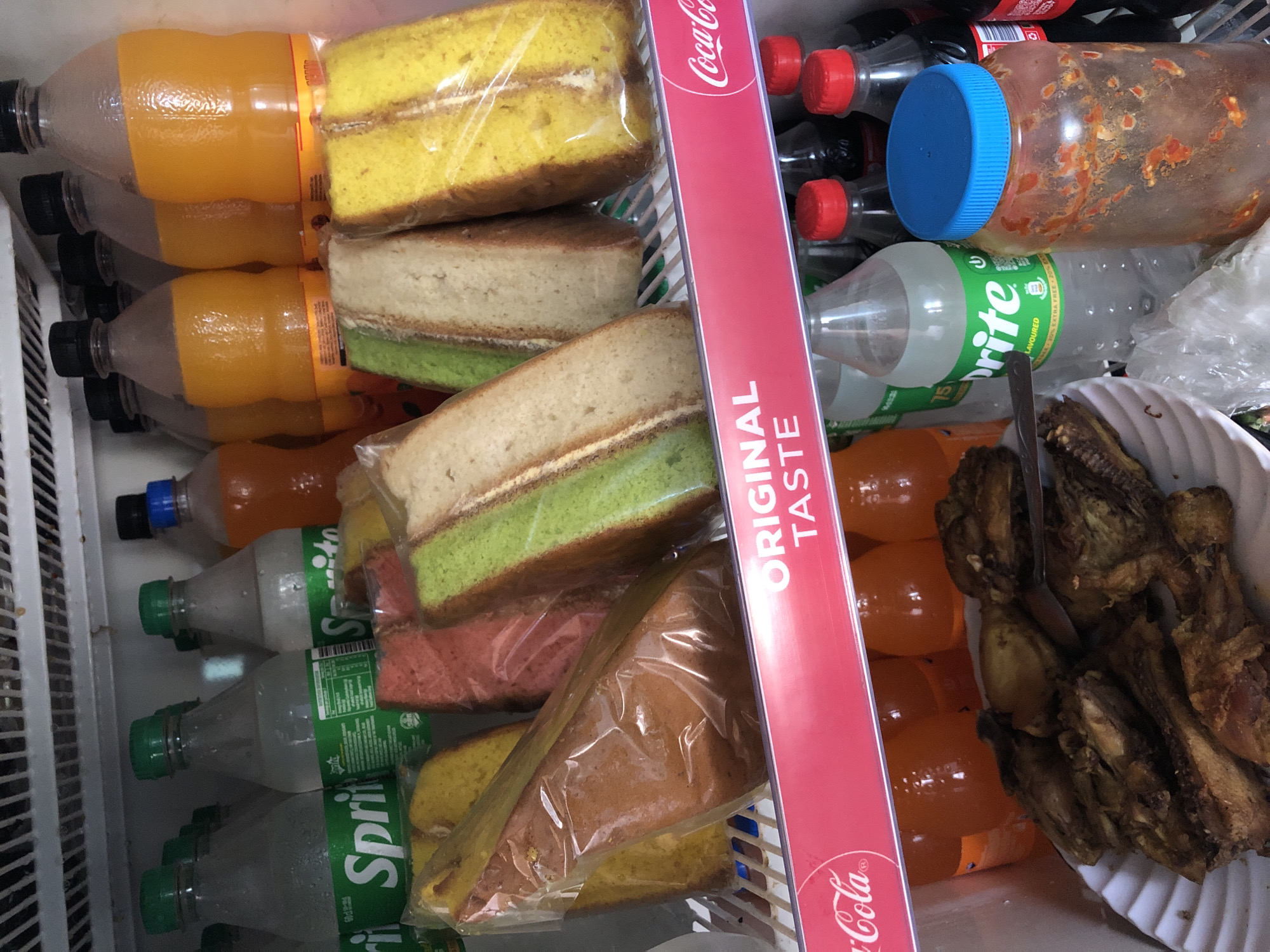 The image shows the inside of a refrigerator, likely in Jos, Nigeria, displaying various food and beverage items. On the top shelf, there are several bottles of orange soda and what appear to be slices of cake wrapped in plastic. The cakes are distinctively colored, with some being pink, green, and yellow. Below this, there are several bottles of Sprite. In the foreground, on the bottom shelf, a white plate holds a portion of fried chicken pieces. To the right of the chicken, a large, transparent jar with a blue lid is filled with a reddish, chunky condiment, possibly chili paste or sauce. A red Coca-Cola branded shelf divider runs horizontally across the middle of the image, separating the upper and lower contents of the refrigerator. The overall impression is of a domestic or small-scale food storage environment.
