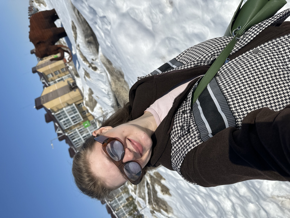 A young woman is captured in a close-up selfie, positioned in the foreground and smiling directly at the camera. She wears brown-framed reflective sunglasses, a dark brown hooded jacket, a houndstooth-patterned vest with black and white horizontal stripes across the chest, and a light pink undershirt. A dark green shoulder bag is visible across her body. The ground is covered in significant amounts of white and partially melted, dirty snow. In the middle ground, to the right of the woman, a large, brown sculpture of a woolly mammoth with tusks stands on a snow mound. The background features several multi-story residential buildings with beige facades, multiple windows, and dark roofs, under a clear blue sky. A street light and a partially visible green sign are also present near the buildings. The scene is located in Новое Шигалеево, Russia.